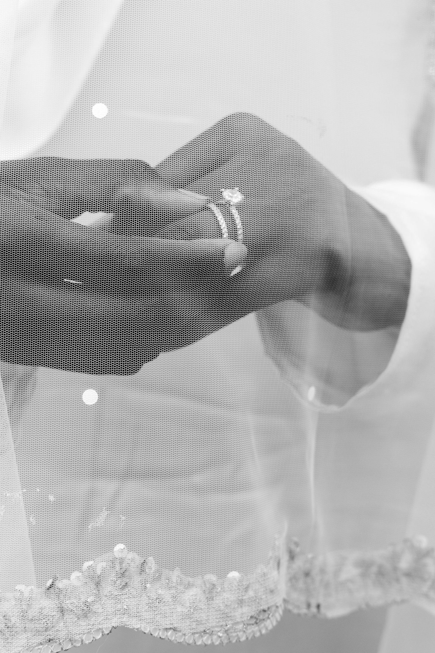 Black and white close-up of hand wearing engagement ring with lace veil in background. | PHL, PA Wedding Photographer | Desi Weddings