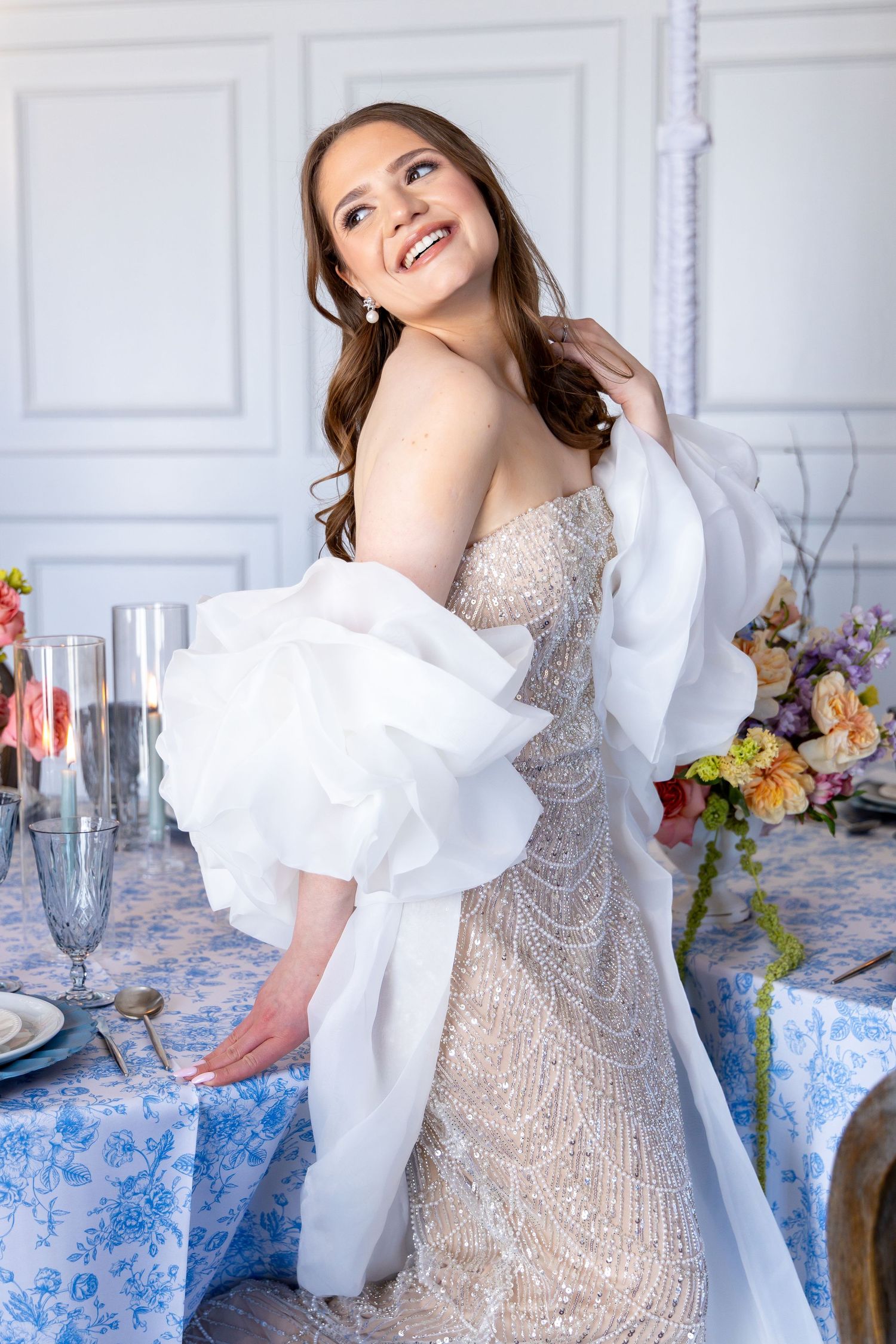 Bride in beaded gown and white shawl smiles during elegant wedding photoshoot with floral decor and table settings.