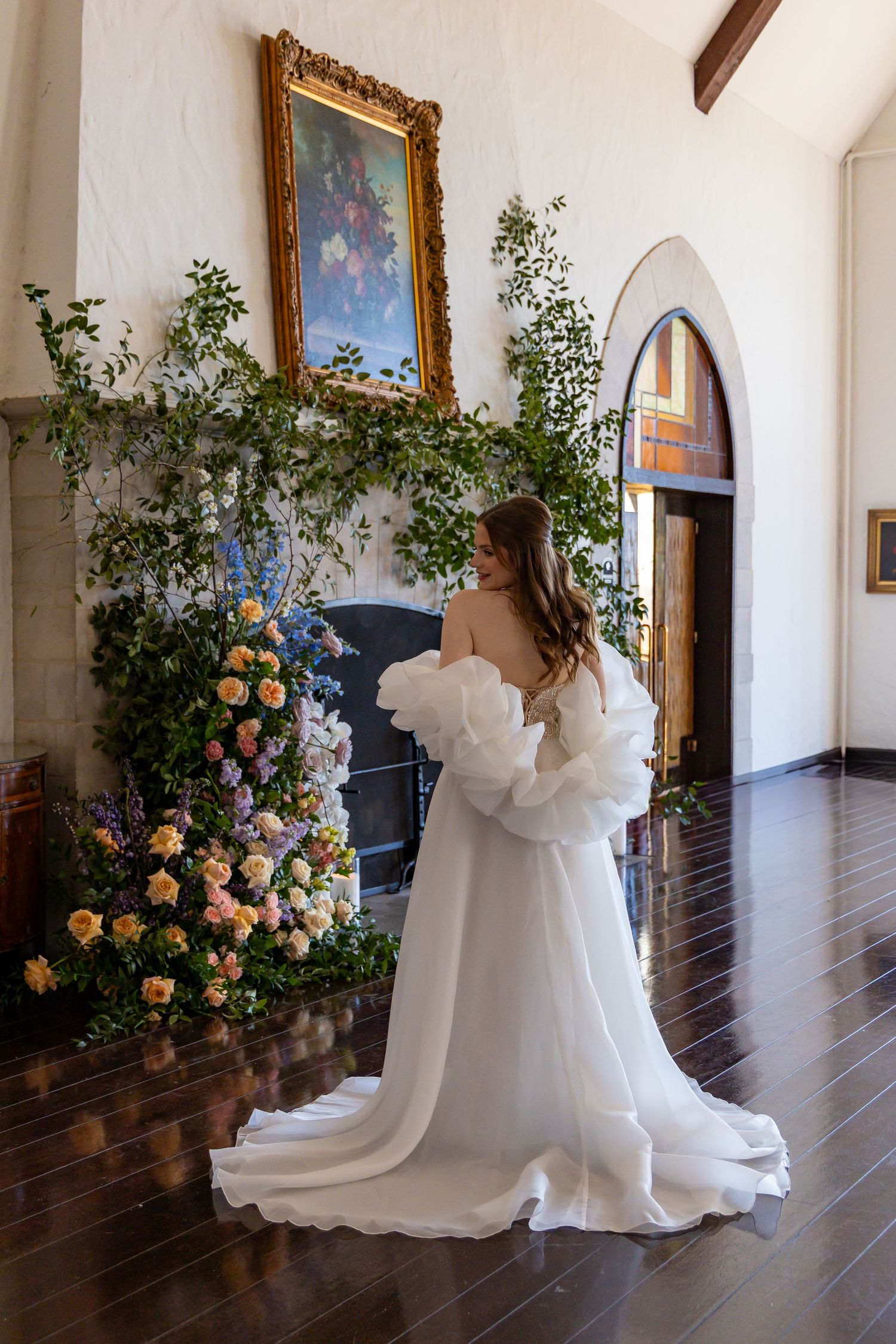 Wedding gown with flowing train photographed against an elegant interior with floral arrangements.