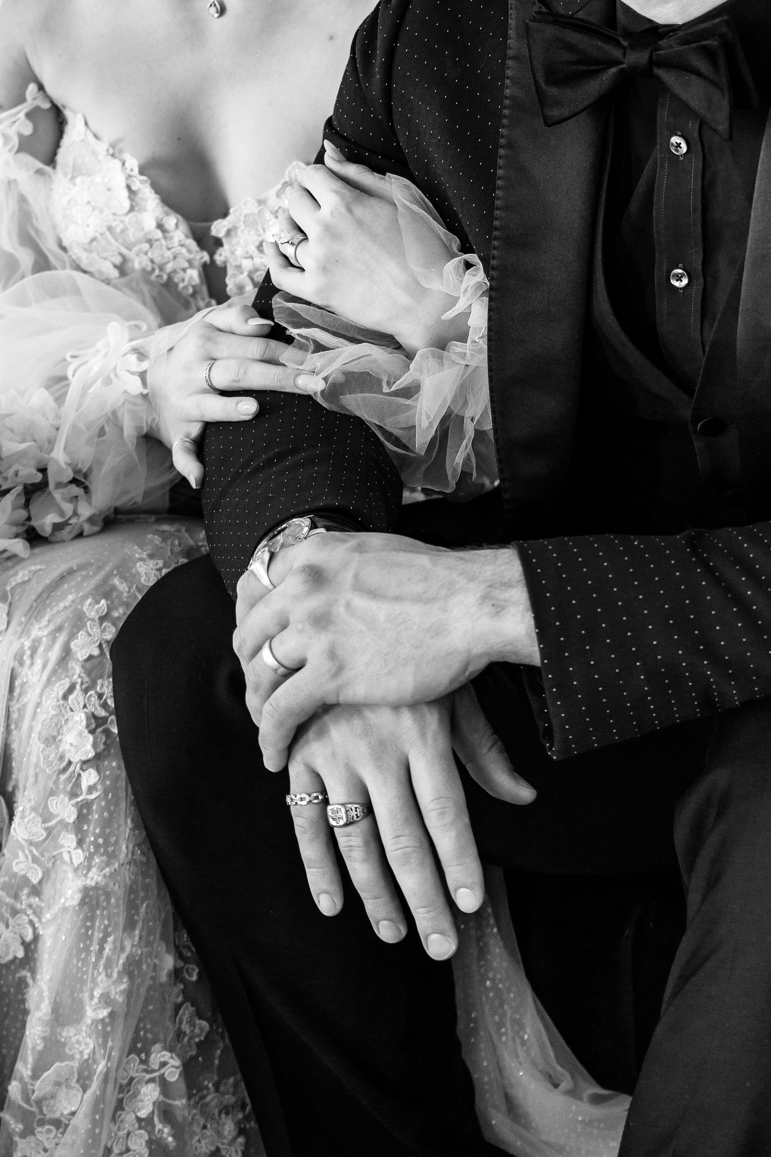 Black and white close-up of hands with wedding rings intertwined against formal wedding attire.