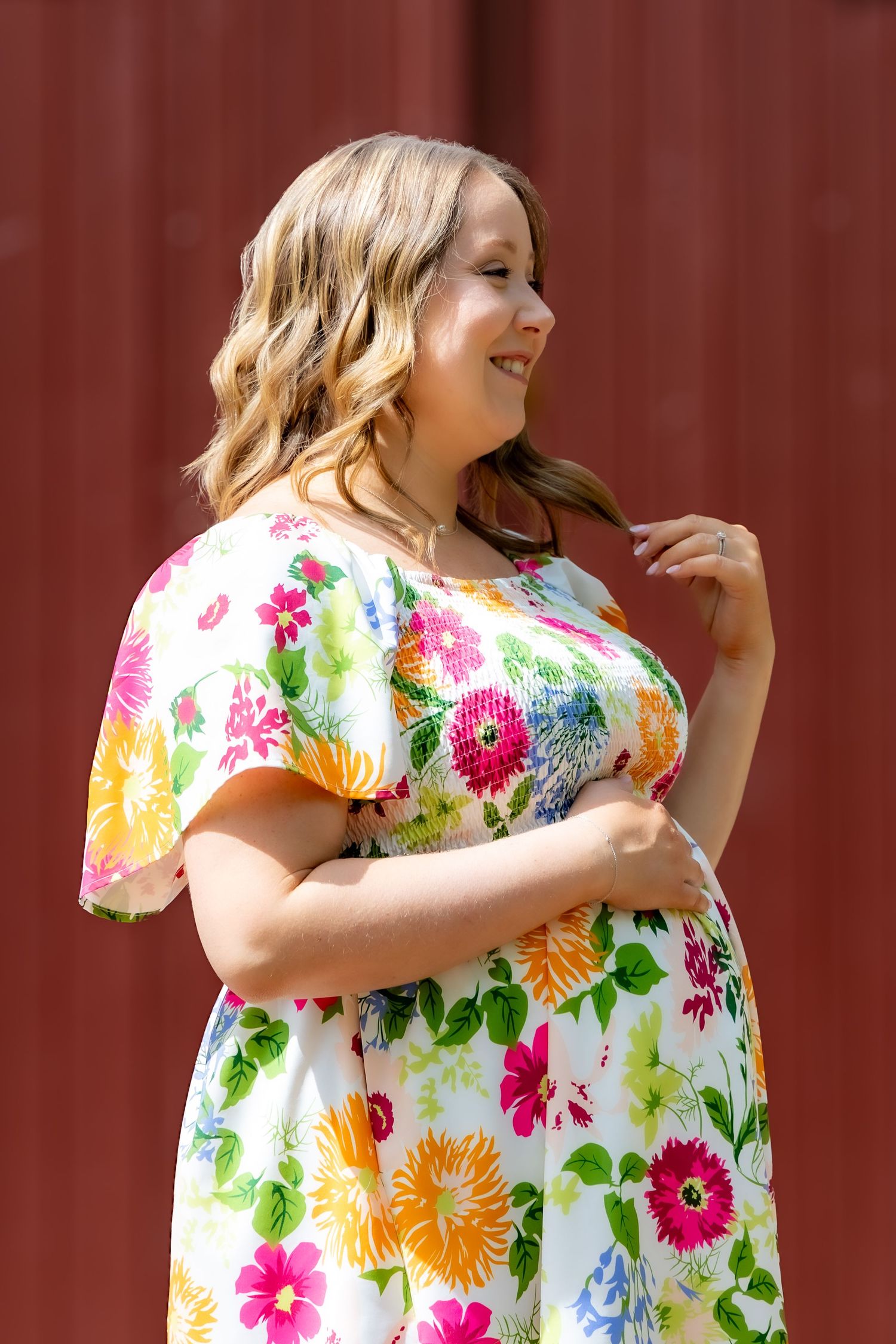 Woman in a colorful floral dress poses against a red wall outdoors.