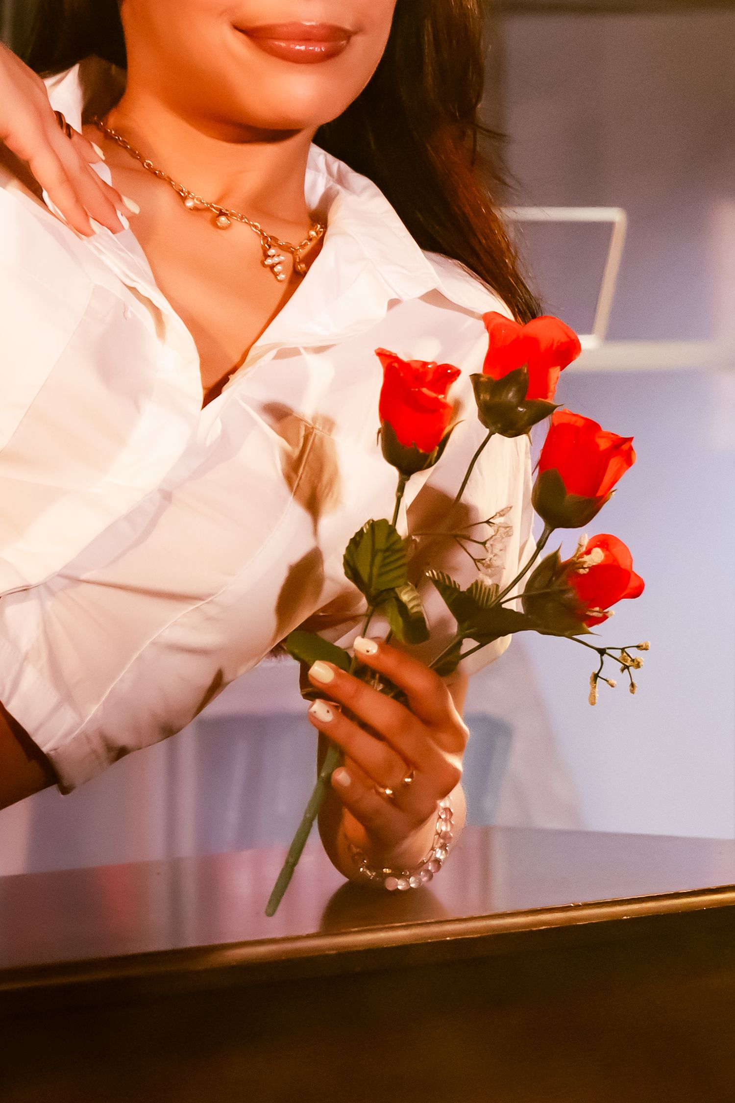 A person wearing a white shirt holds red roses and a delicate gold necklace in a warm-toned vintage style photograph.