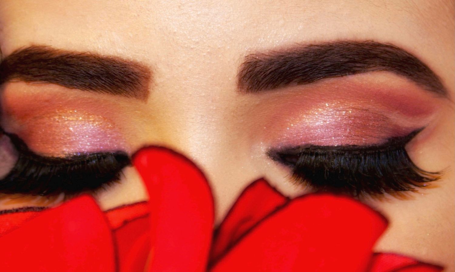 Close up view of eye makeup with pink and rose gold eyeshadow, dramatic winged eyeliner, and false lashes against red fabric.