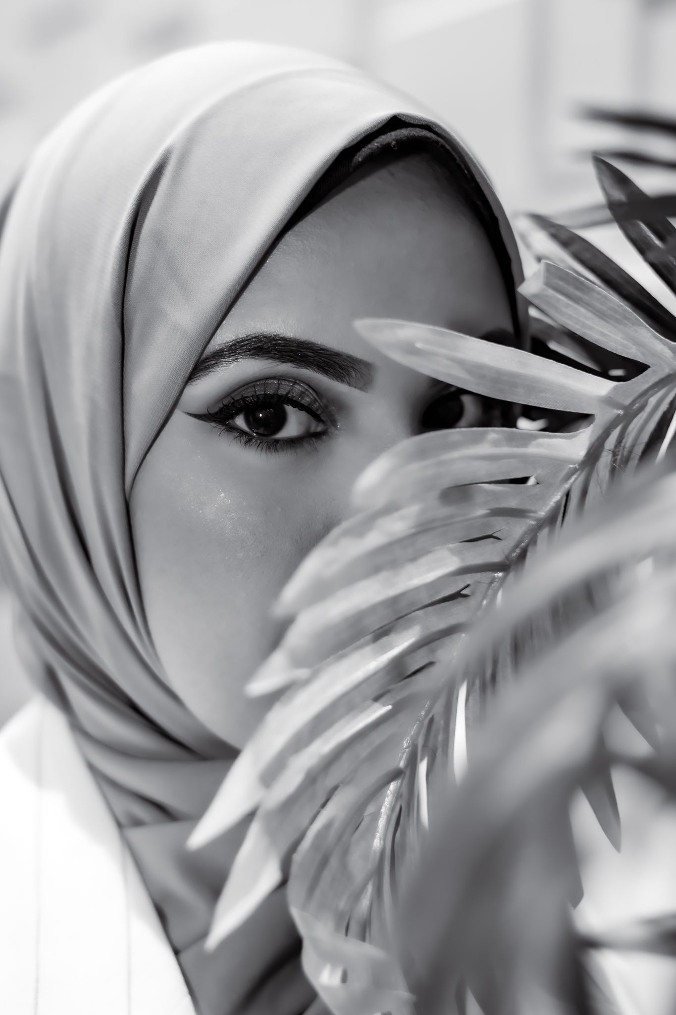 Artistic black and white portrait showing a hijab-covered individual behind palm fronds.