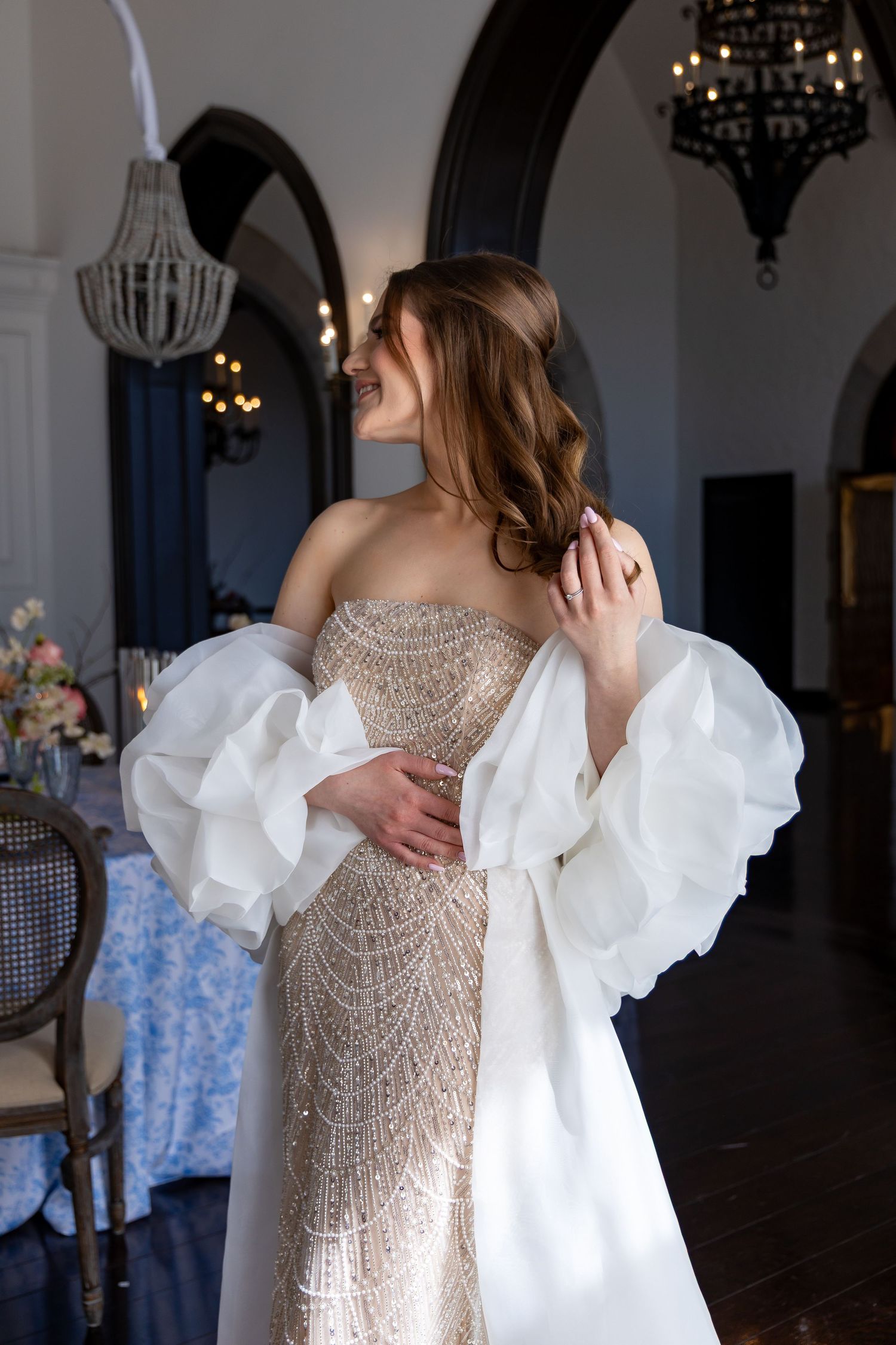 Elegant beaded wedding gown with dramatic white detachable statement sleeves in an ornate venue with chandeliers.