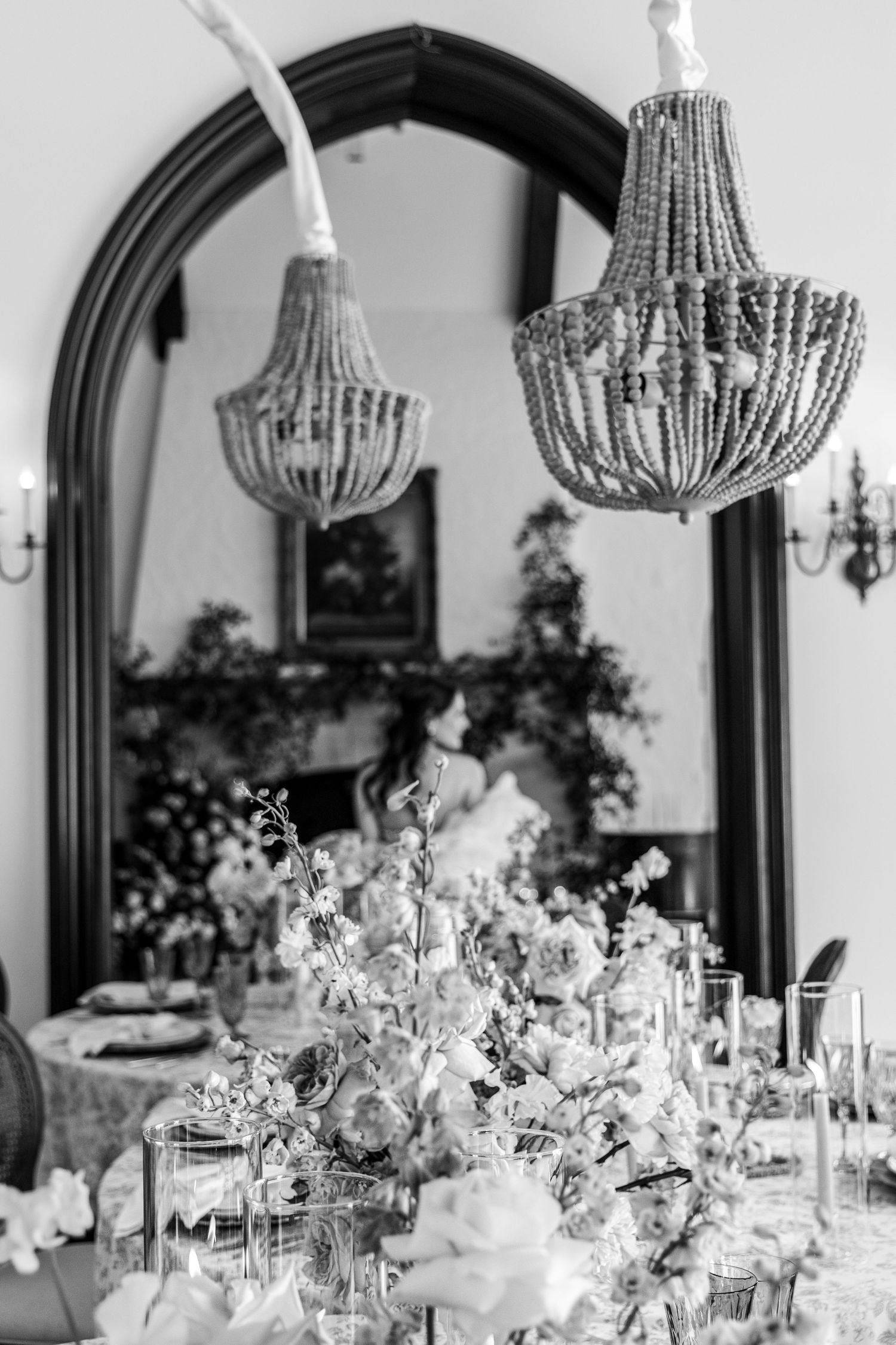 Elegant beaded chandeliers hang above a romantic table setting with floral centerpieces and mirror arch decor.