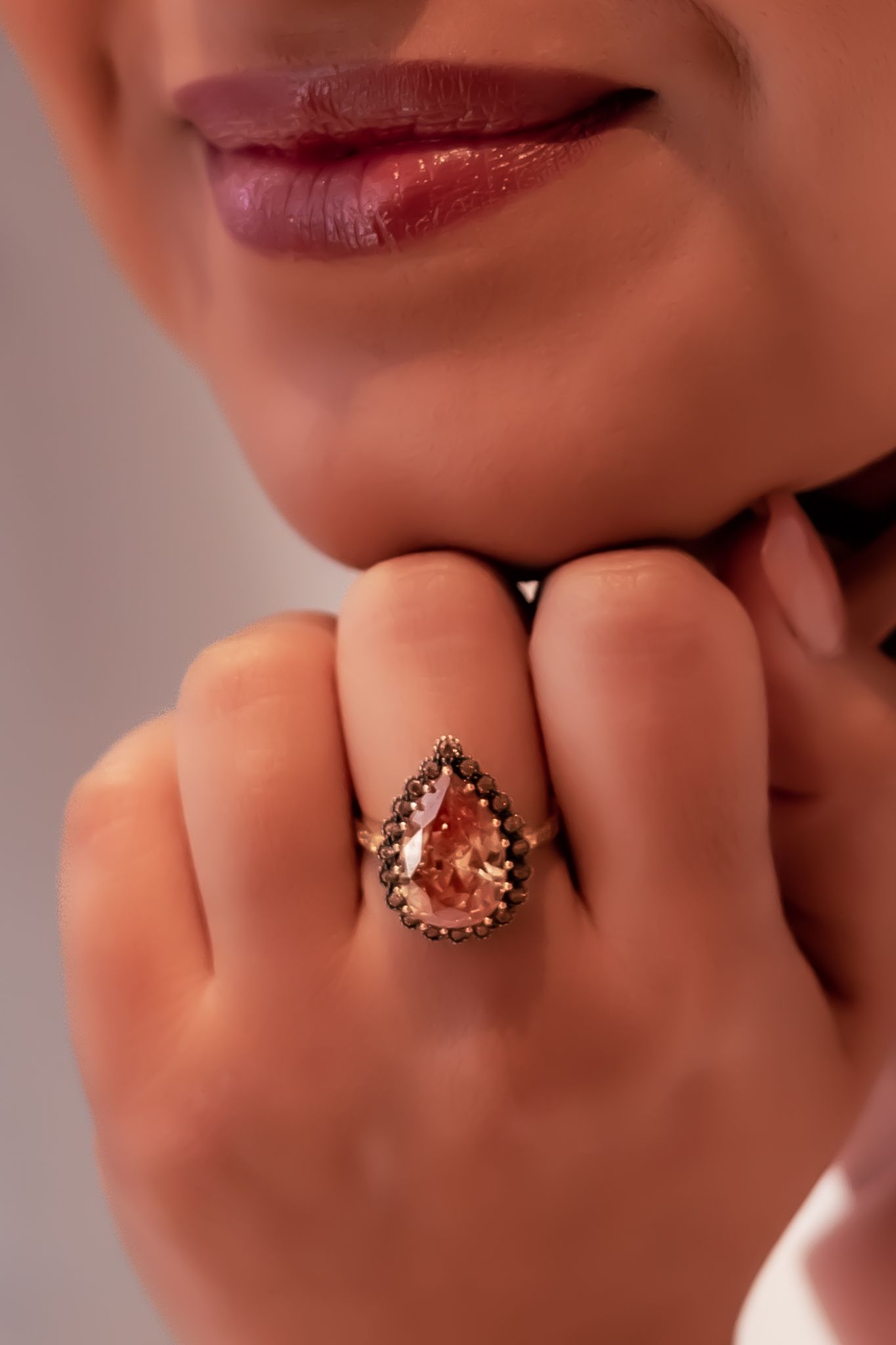Close-up of a hand wearing an elegant teardrop-shaped rose gold ring with dark red lipstick in soft focus background.