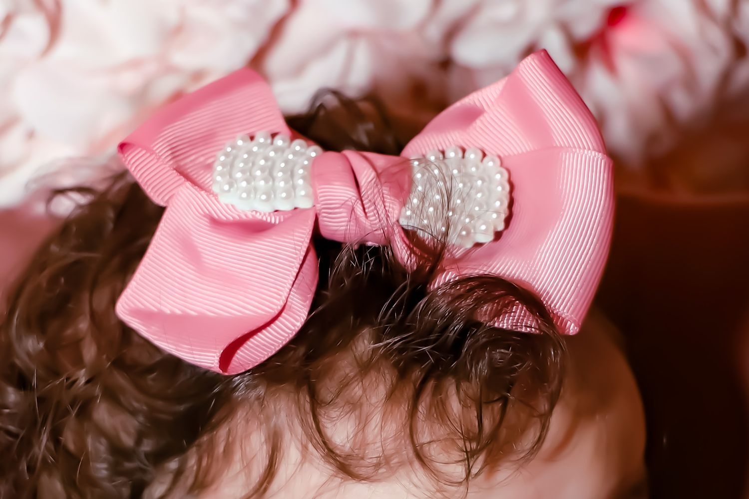A pink satin hair bow with pearl embellishments adorns dark curly hair.