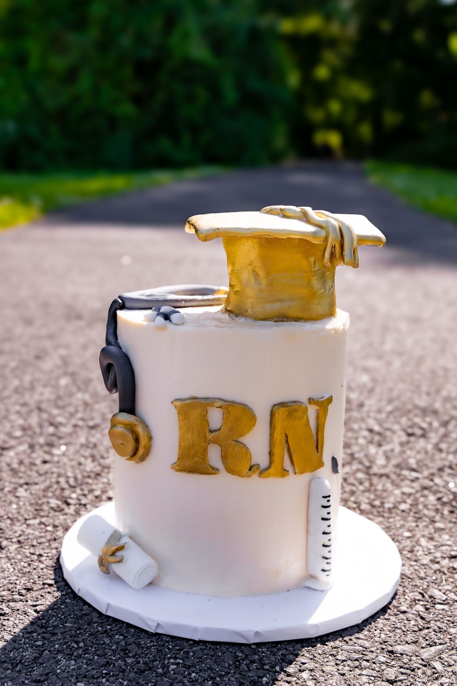 A white fondant cake decorated with gold RN letters and a graduation cap, showcasing a nursing theme on an outdoor path.