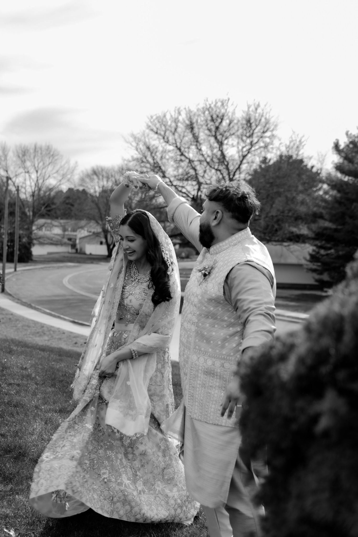 A candid black and white wedding photo shows a couple walking together outdoors on a curved path surrounded by bare trees.