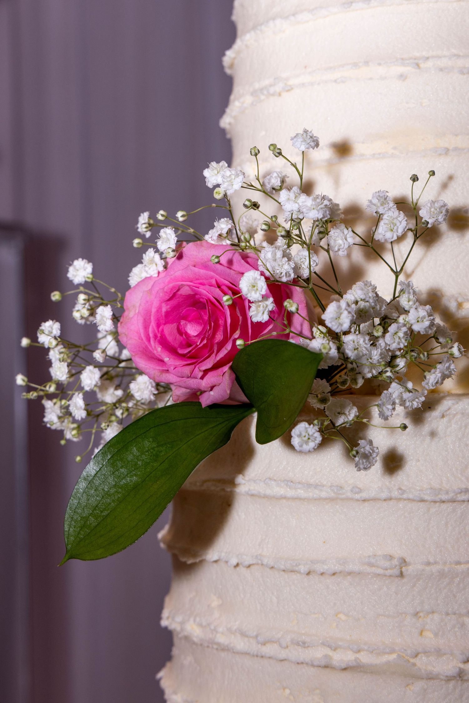 Close-up of a white wedding cake decorated with a pink rose and baby's breath flowers against a purple background.