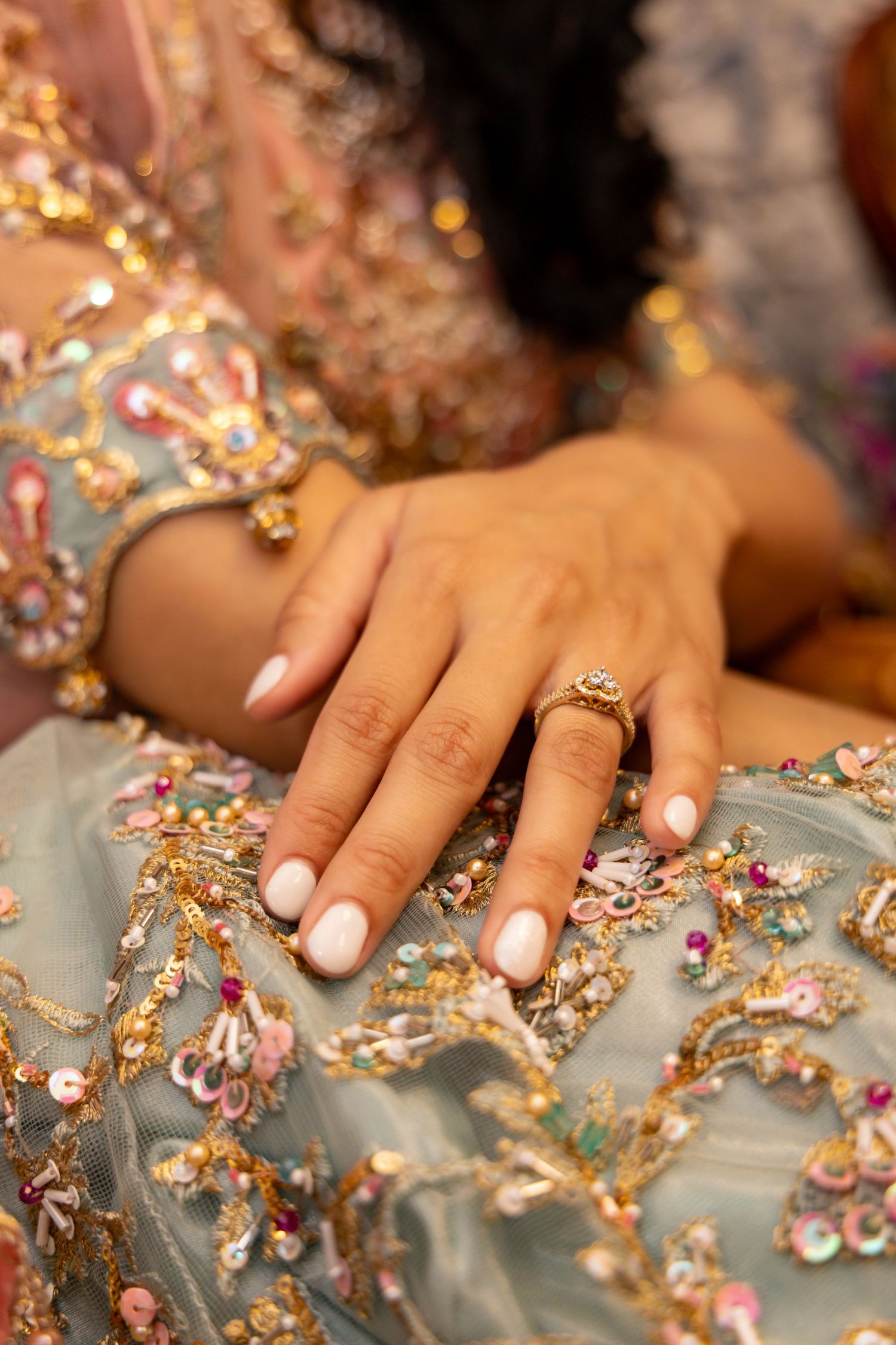 Close up of a hand wearing French manicure and engagement ring against an embellished light blue gown with golden details.