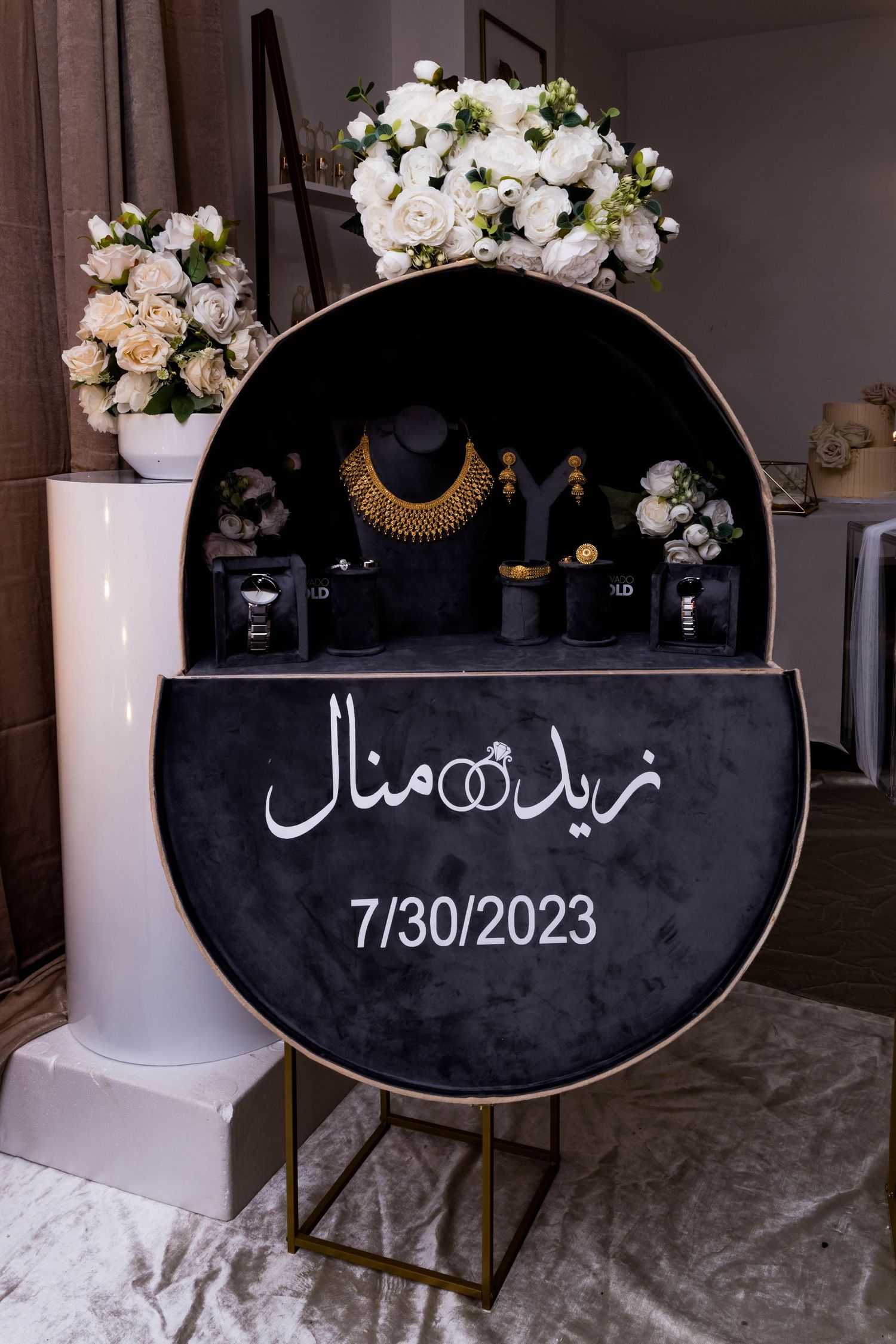 Wedding decor display featuring black circular sign with Arabic text and date 7/30/2023, adorned with white floral arrangements.