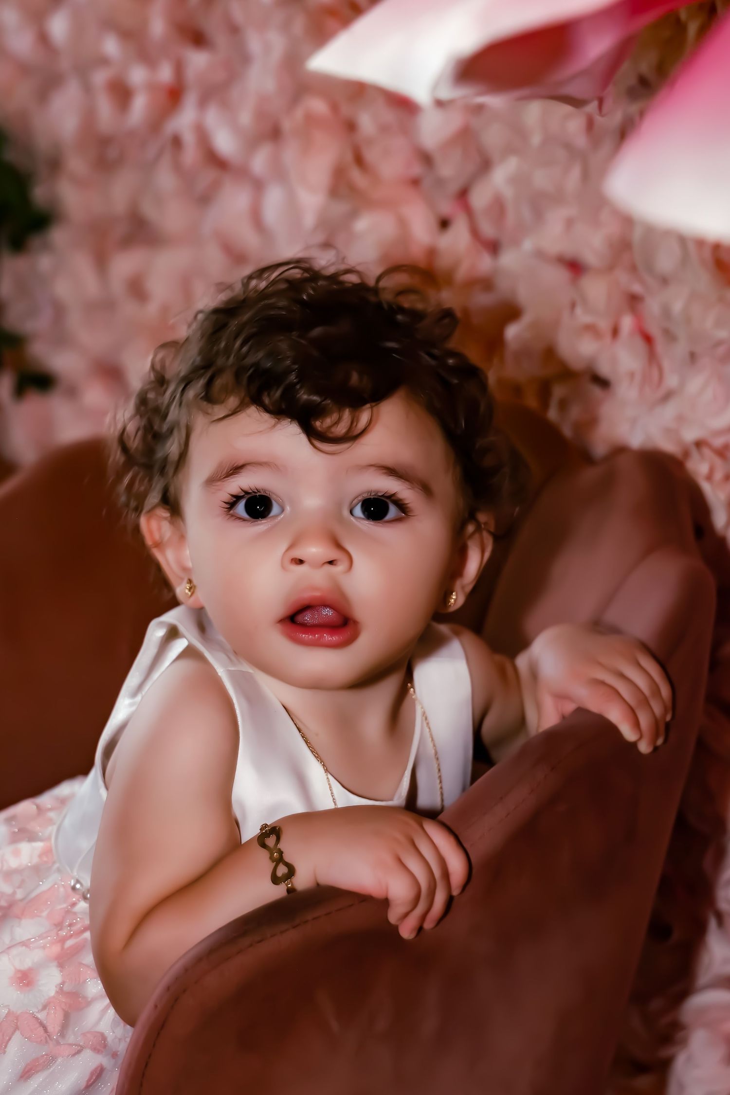 A toddler in a white sleeveless top peeks over a brown couch against a pink floral backdrop.