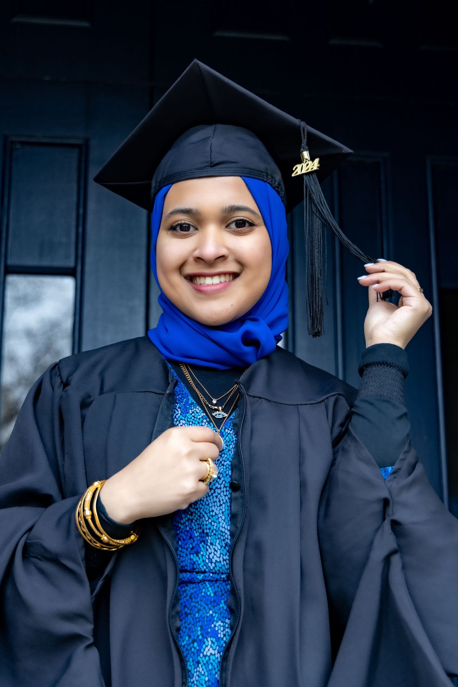 Graduate wearing blue hijab and traditional black graduation gown and cap poses with cheerful expression during ceremony.