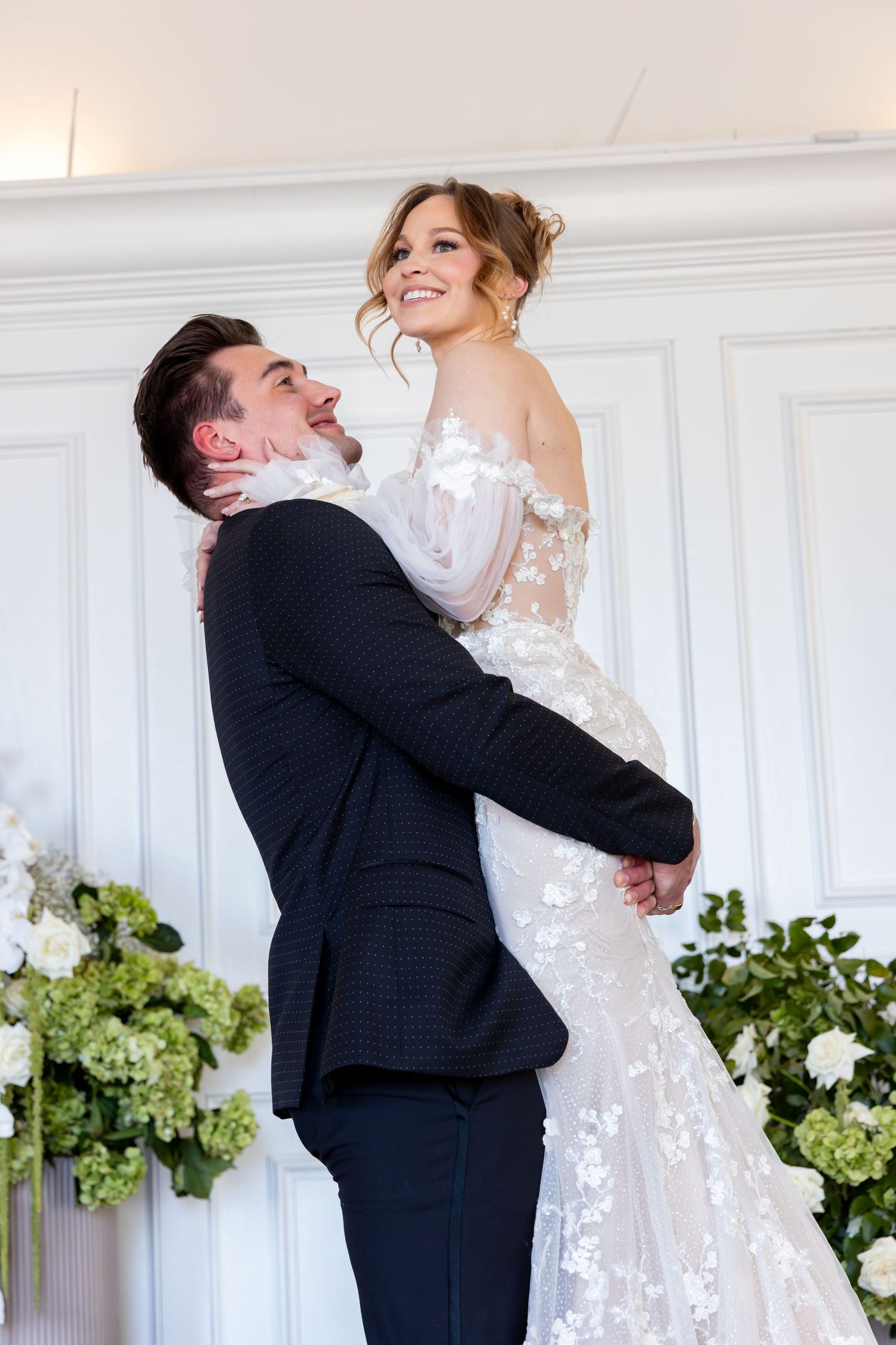 Playful wedding photo of groom lifting bride in front of white wall with floral arrangements.