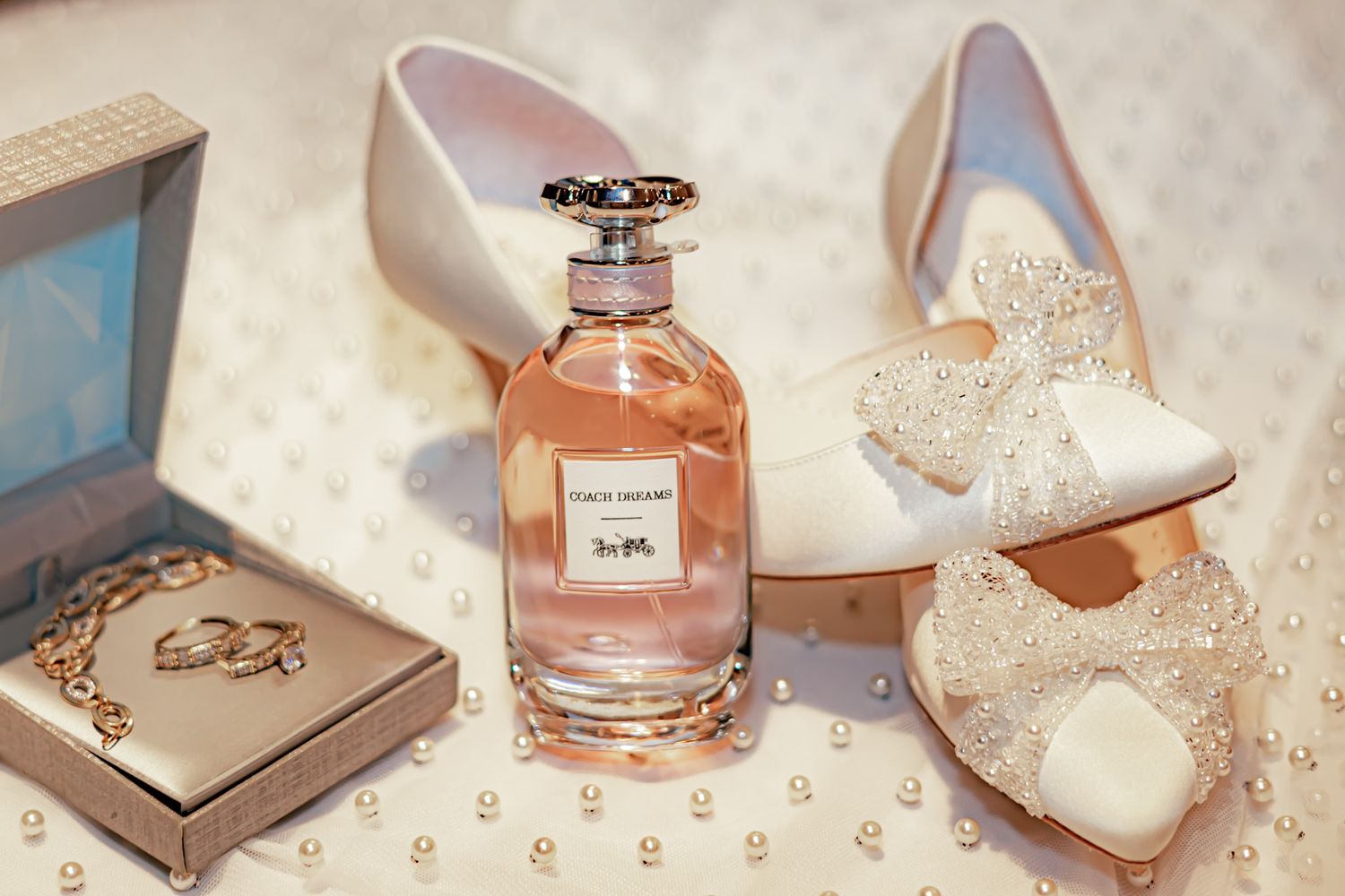 Elegant wedding accessories including white lace heels, perfume bottle, and jewelry box arranged on soft fabric.