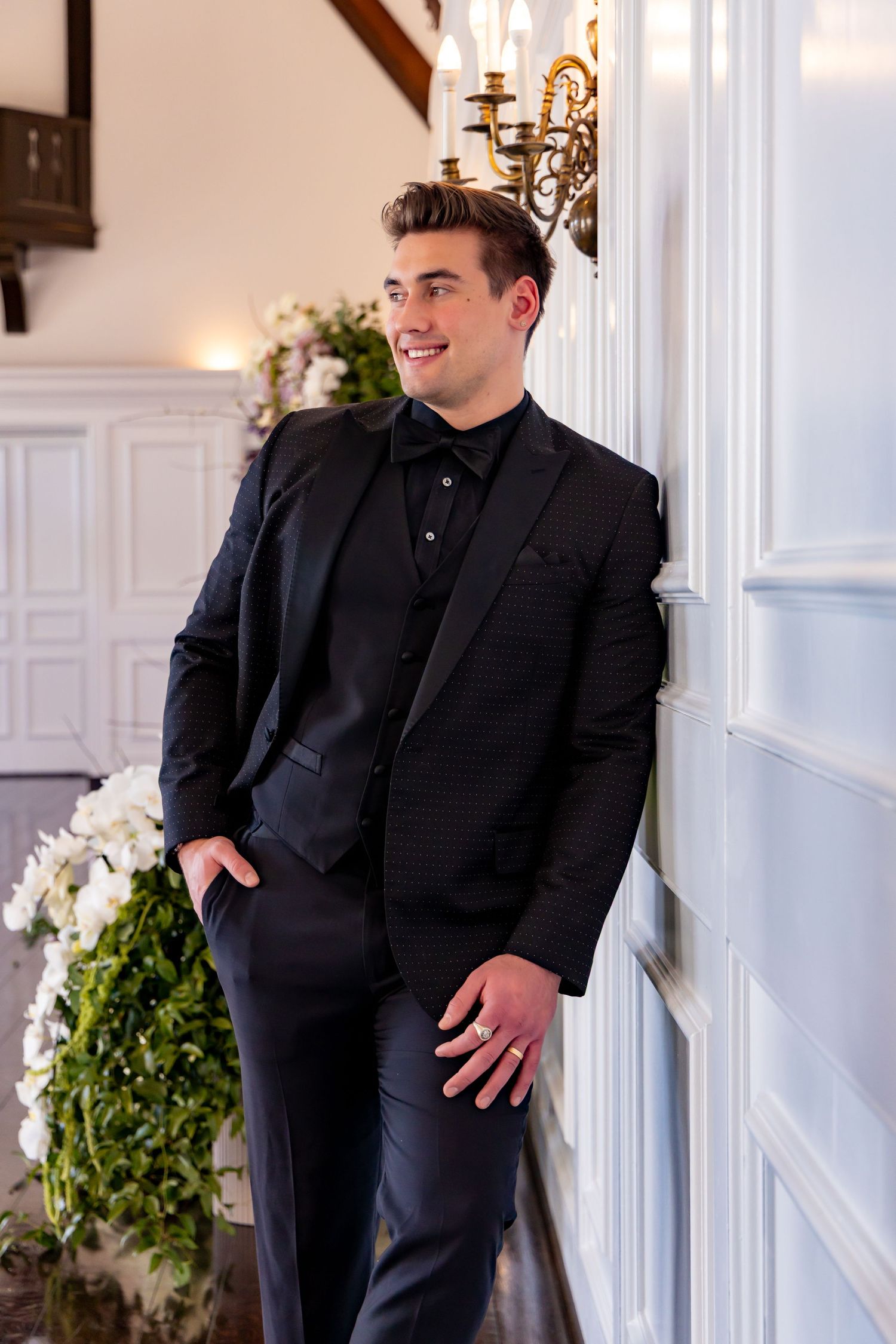 A person in a black formal suit and tie leans against a white wall with elegant decor and floral arrangements.