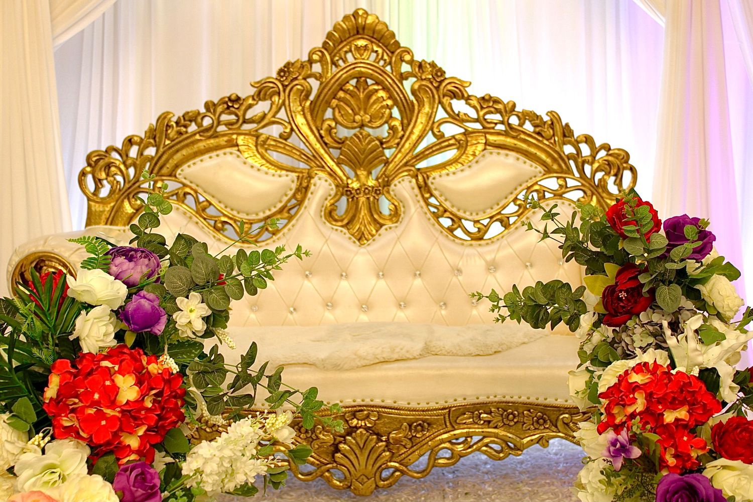 Ornate golden throne with tufted white upholstery decorated with colorful floral arrangements and dramatic lighting.