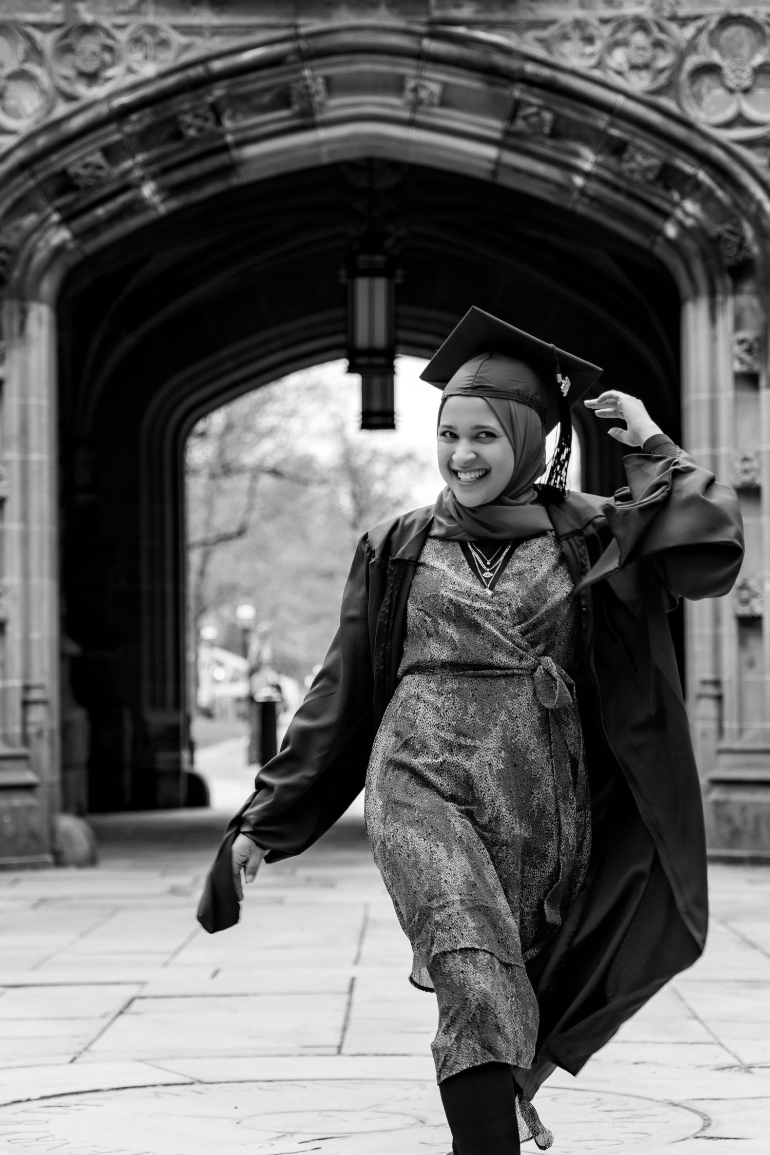 A graduate in cap and gown celebrates joyfully under an ornate Gothic archway in black and white.