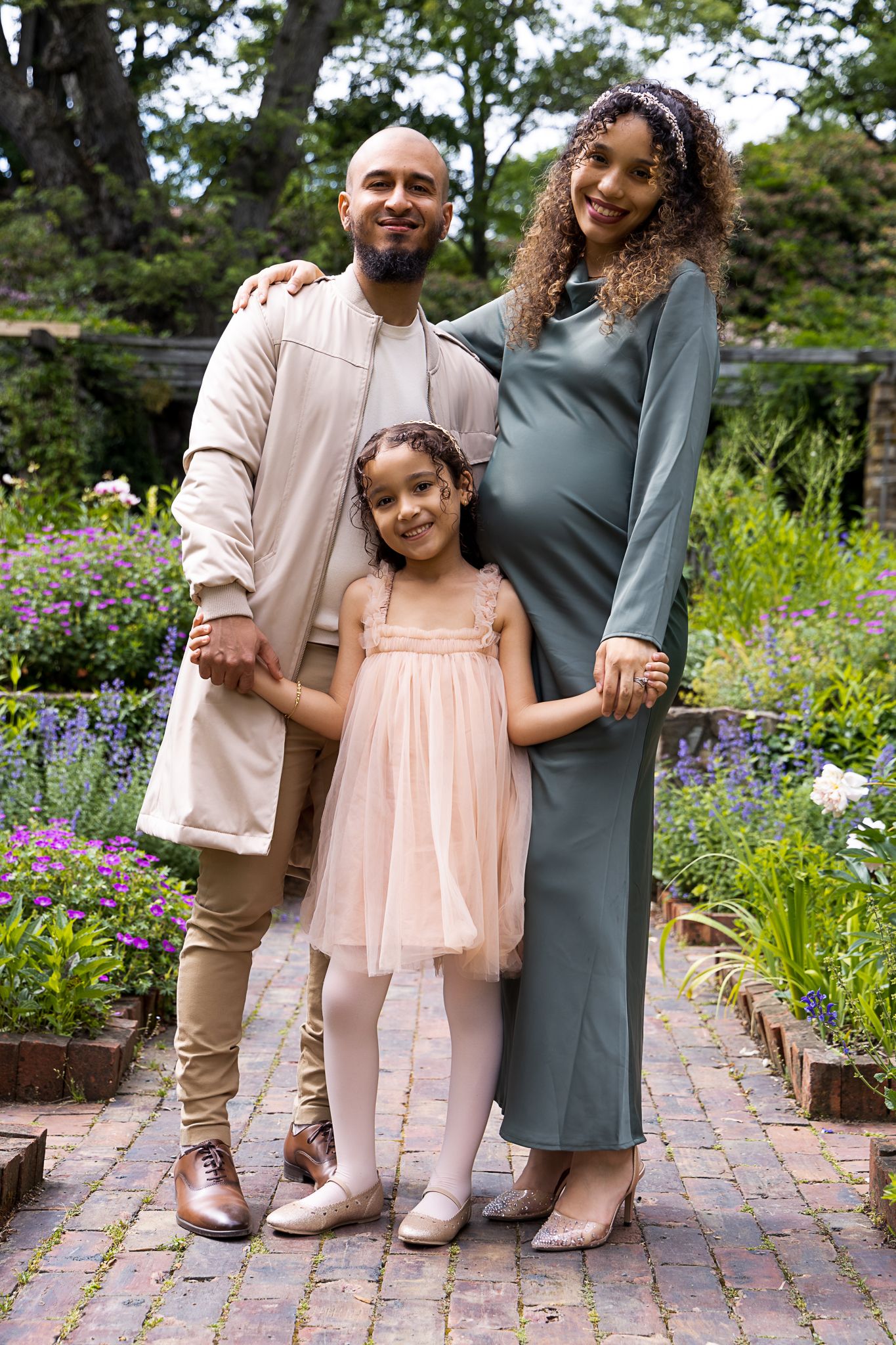 A family of three poses in a garden with blooming flowers, wearing coordinated blush pink and gray outfits.