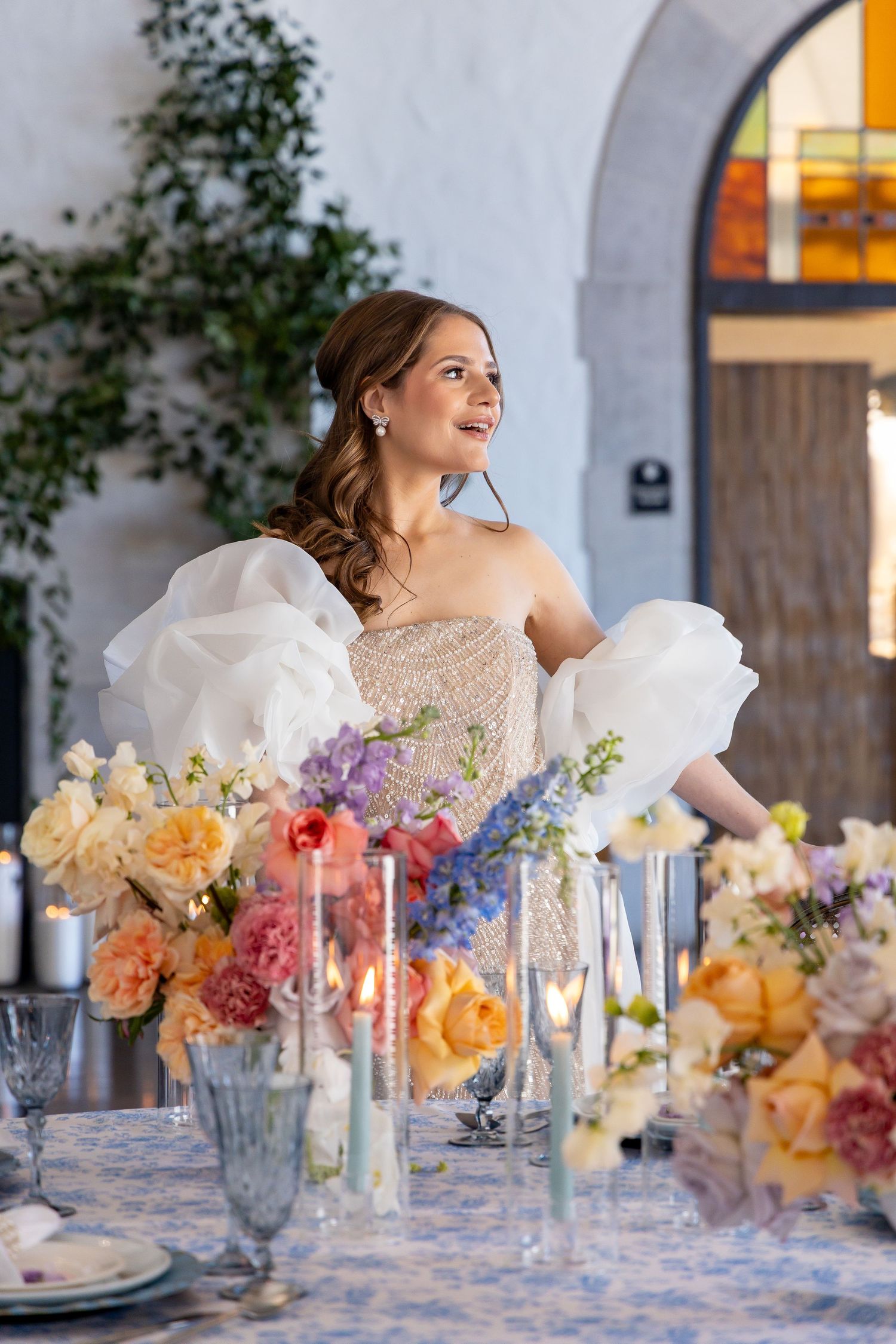 Bride in off-shoulder gown seated at elegant table with colorful floral centerpieces.