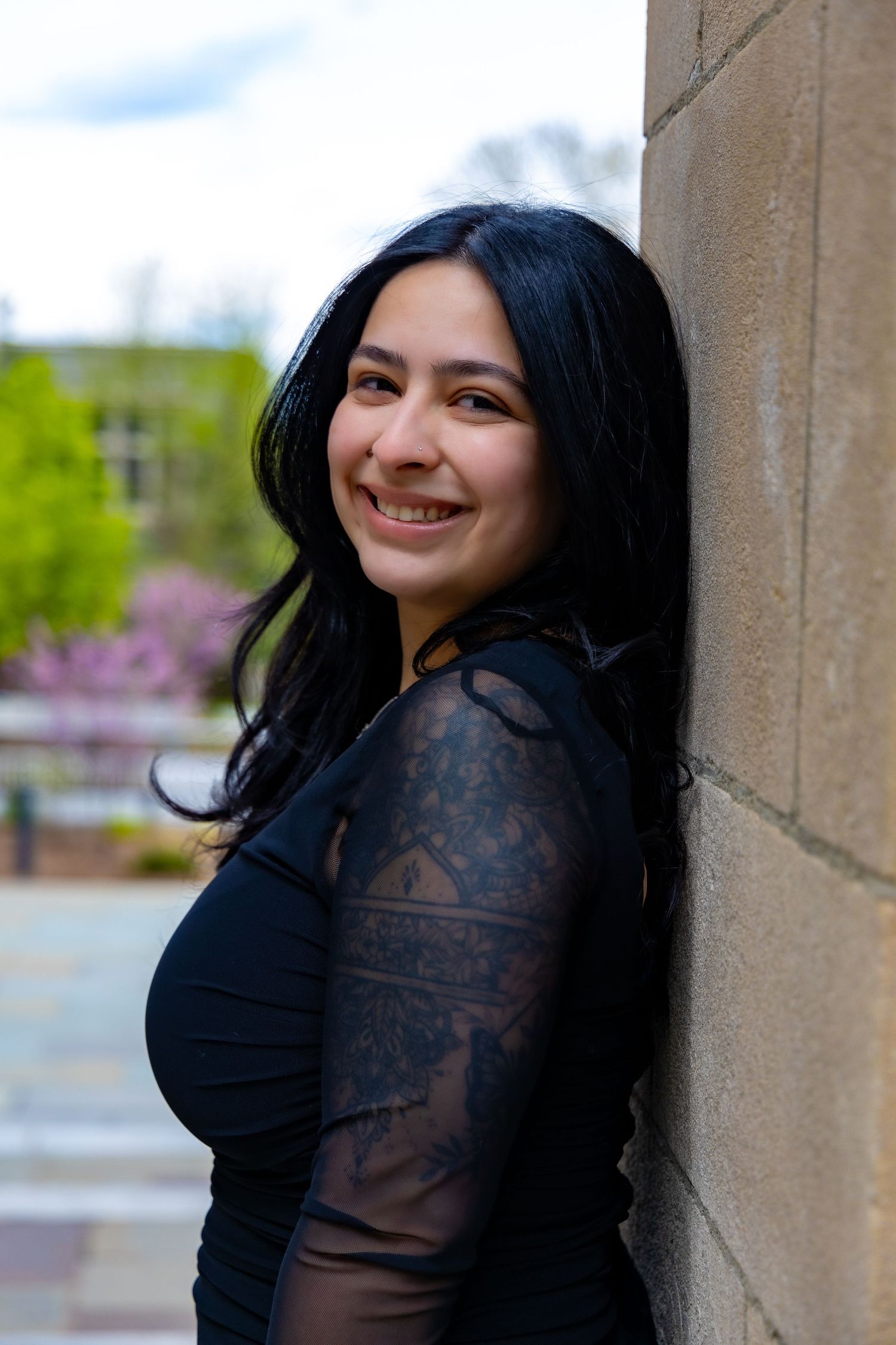 Portrait photo of someone in a black lace top smiling while leaning against a stone wall outdoors.