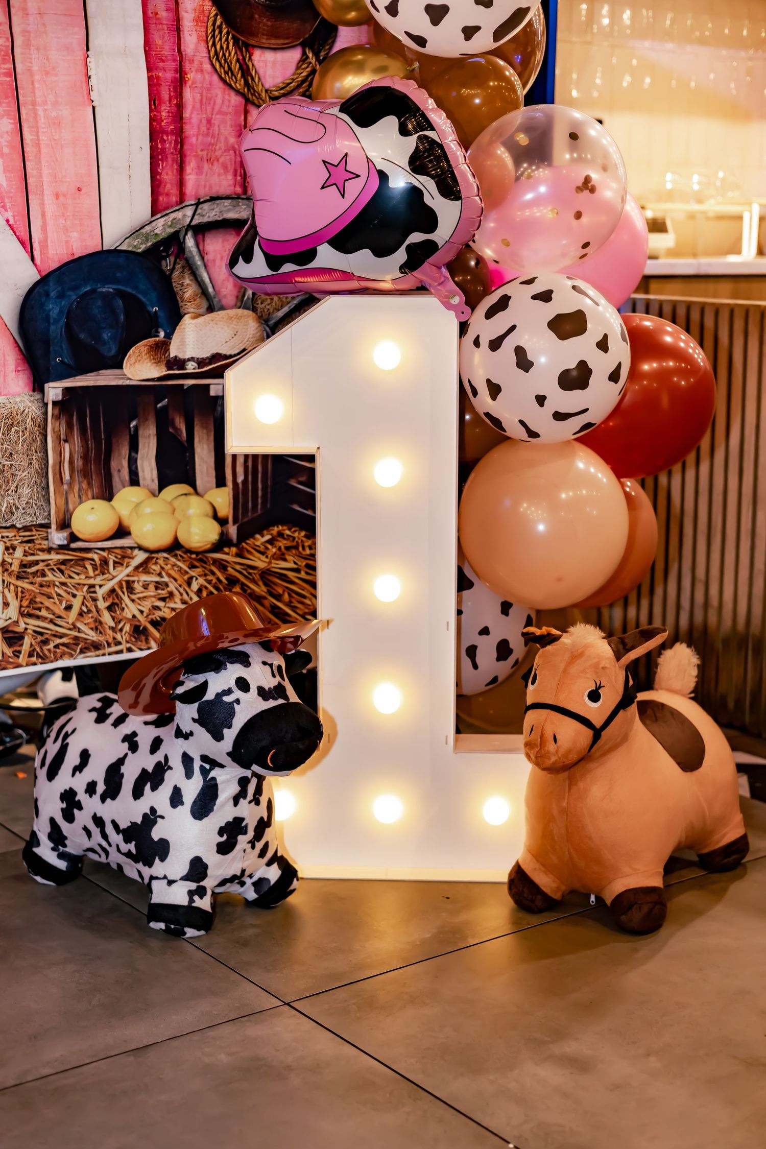 A farm-themed first birthday party display with a light-up number 1, plush animals, and cow print balloons.