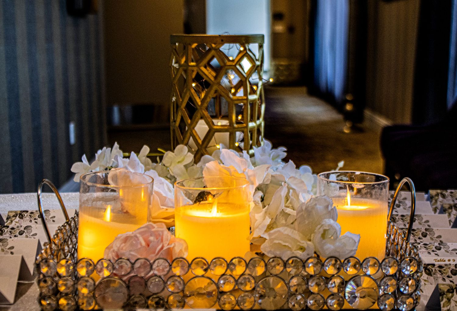 Glowing orange candles in glass holders surrounded by white tulle and gold decorative lanterns on an elegant event table.