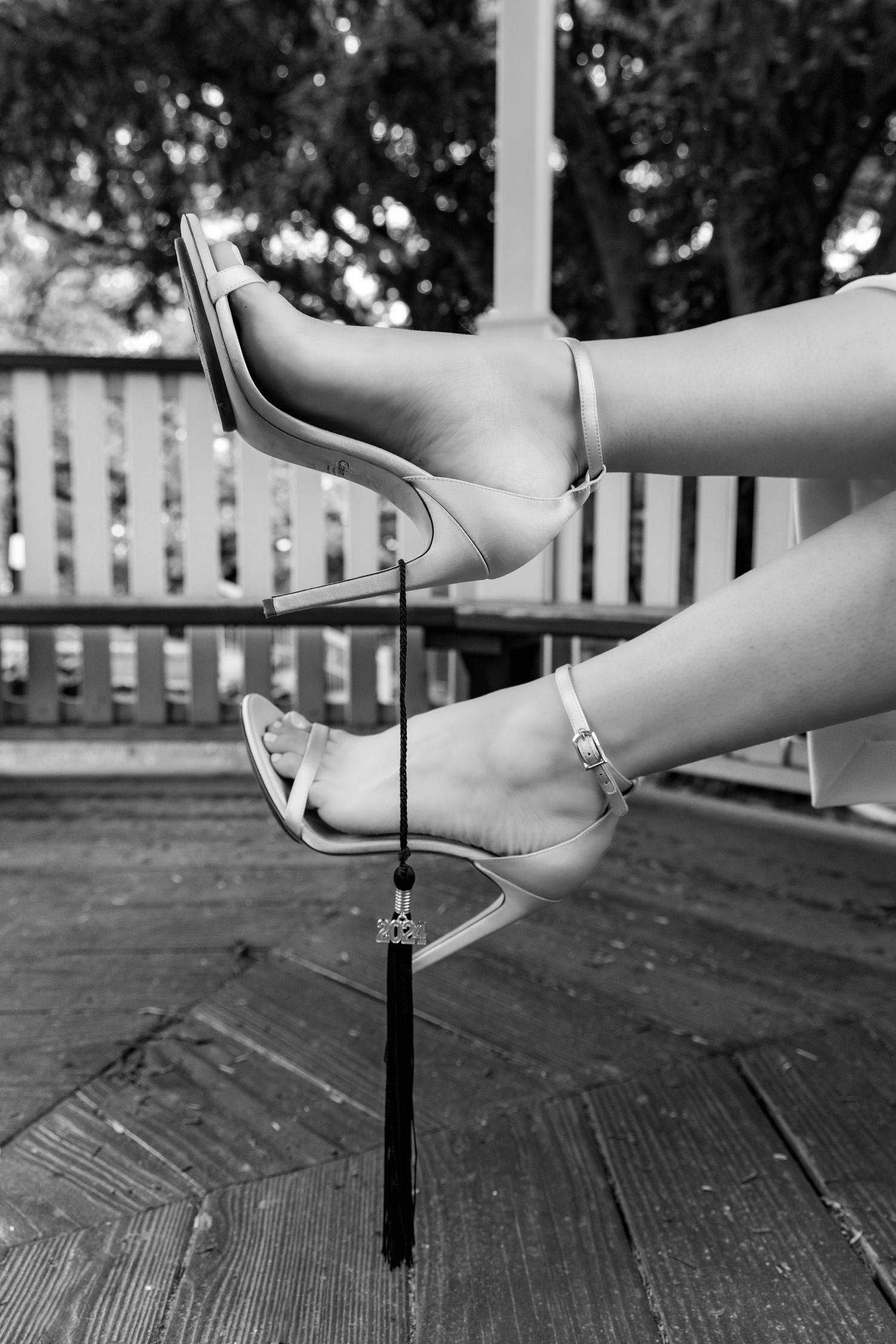 Black and white photo of legs in elegant silver stiletto heels sitting on a wooden deck with a picket fence background.