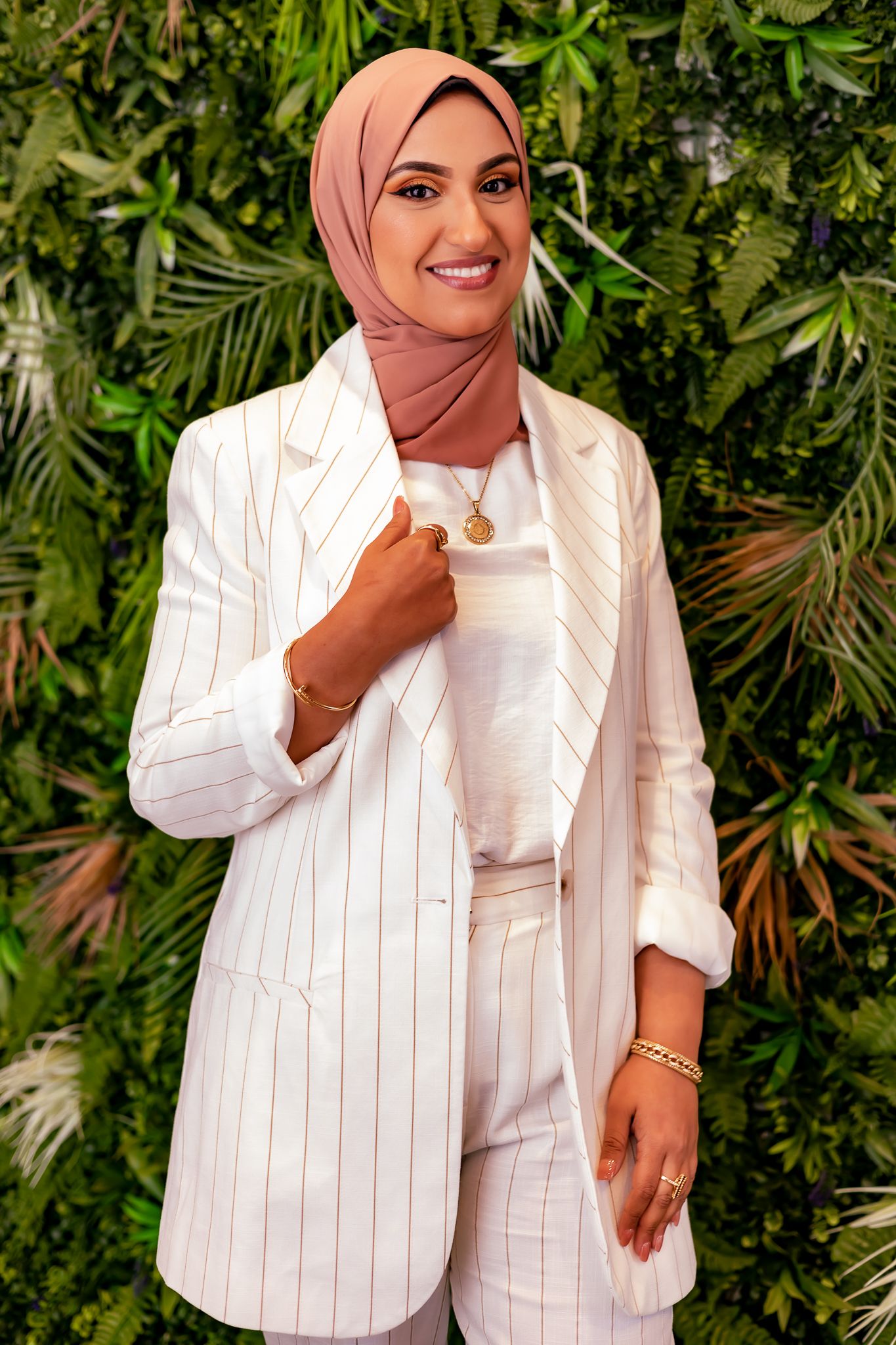 Stylish hijabi wearing a white pinstriped blazer and pants ensemble poses against a lush green tropical plant backdrop.