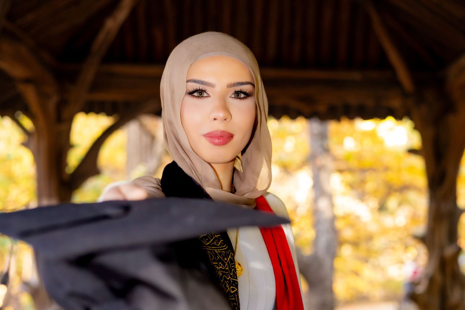 A woman wearing a hijab and academic graduation attire poses outdoors with warm autumn colors in the background.