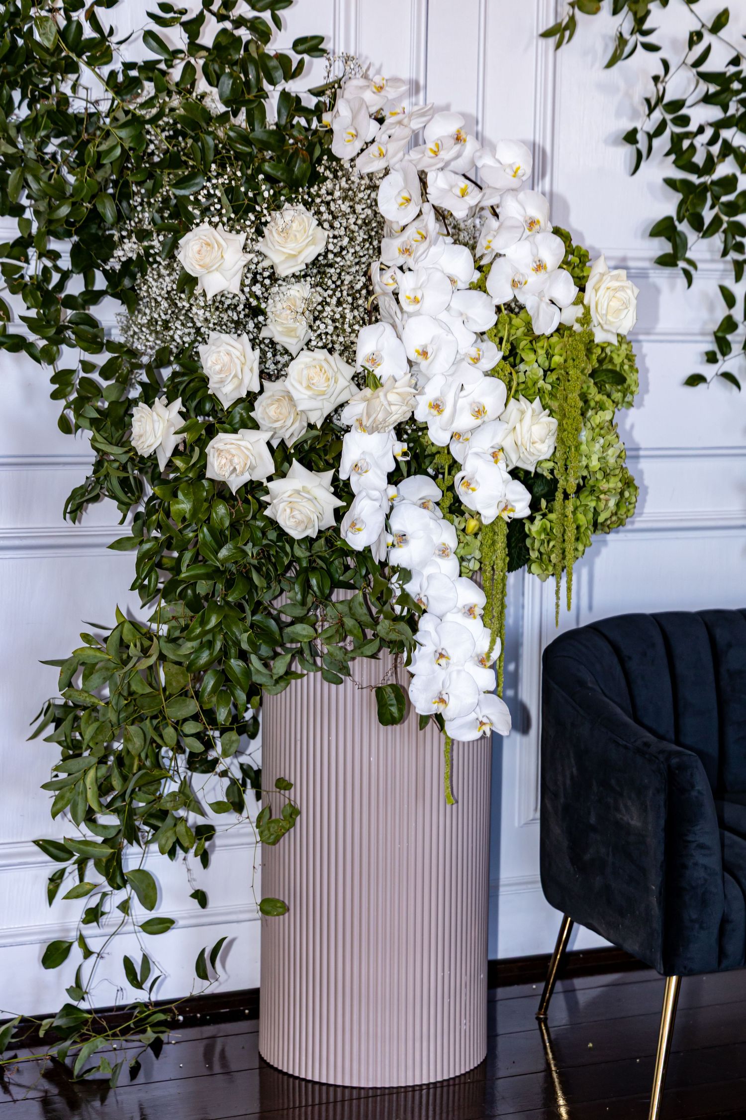 A luxurious floral arrangement of white orchids, roses and baby's breath in a modern gray cylindrical vase against a white wall.