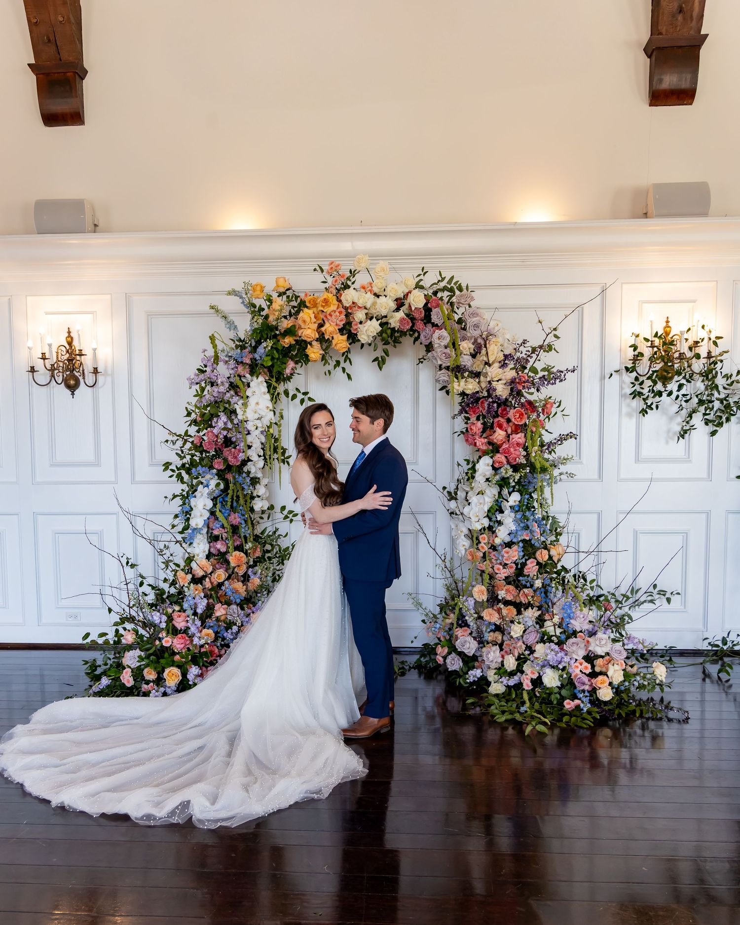 A romantic wedding ceremony setup with an elegant floral arch featuring pink, yellow and purple blooms against white paneled walls.