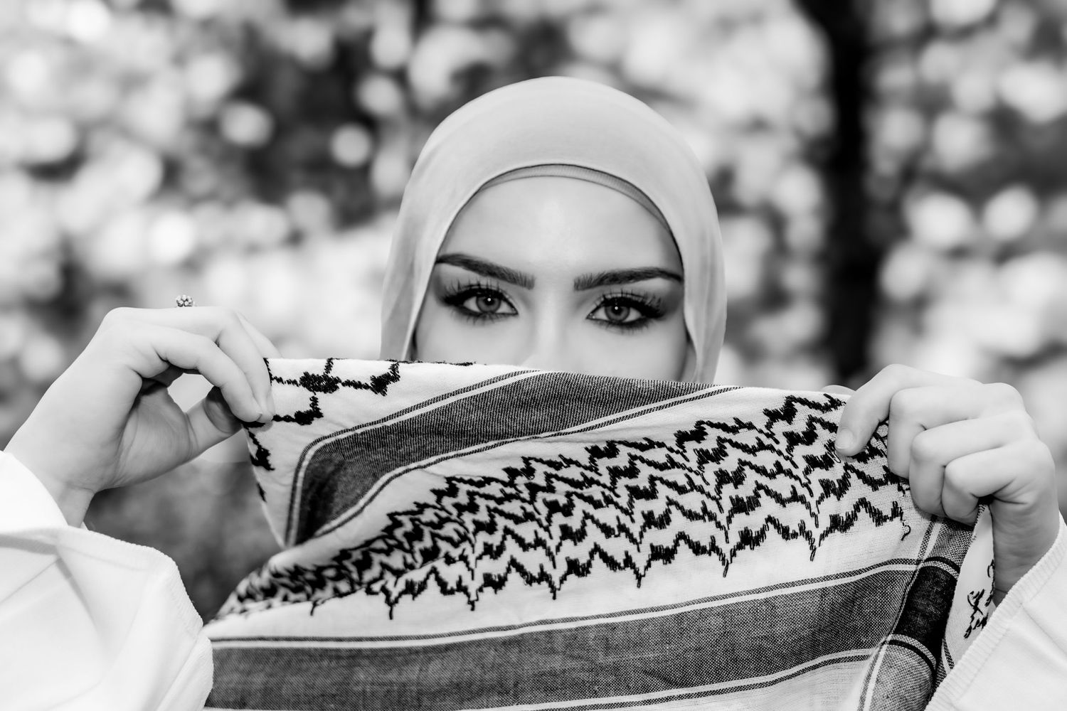 A person wearing a white hijab holds up a patterned scarf with geometric designs in black and white photography.