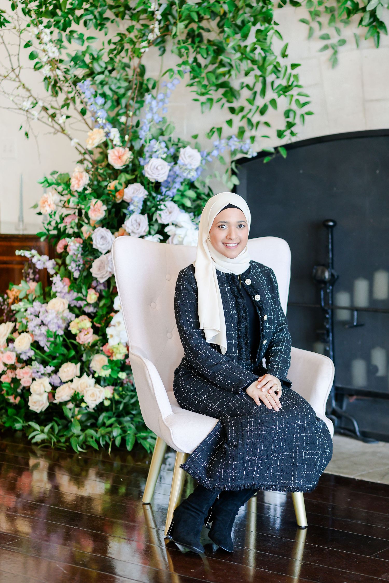 A woman in a white hijab and tweed dress sits elegantly in a white chair surrounded by floral arrangements.
