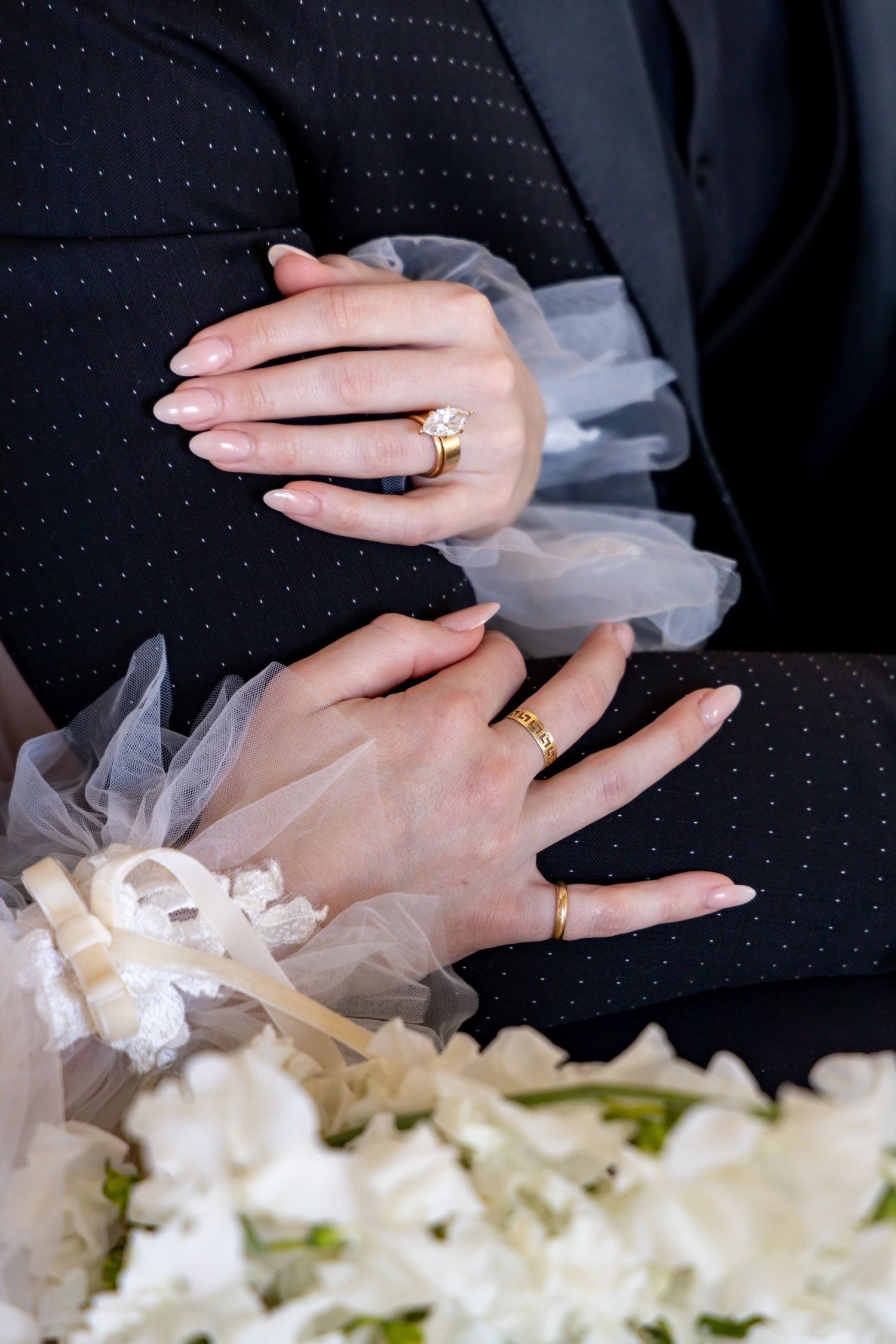 Close-up of hands wearing wedding rings against dark suit with white tulle and floral bouquet details.