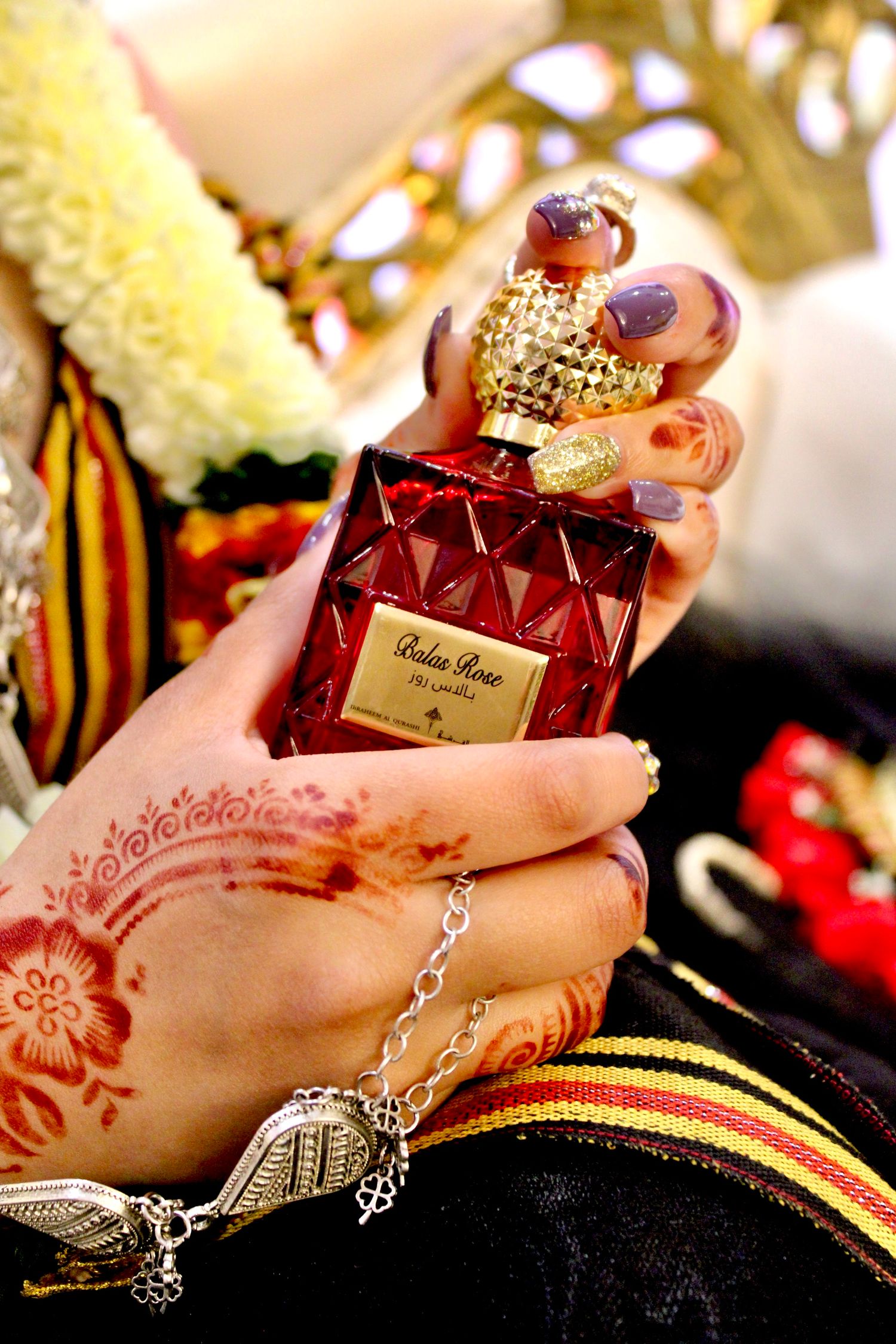 Hand with intricate henna design holds ornate red perfume bottle with gold cap against traditional clothing and jewelry.