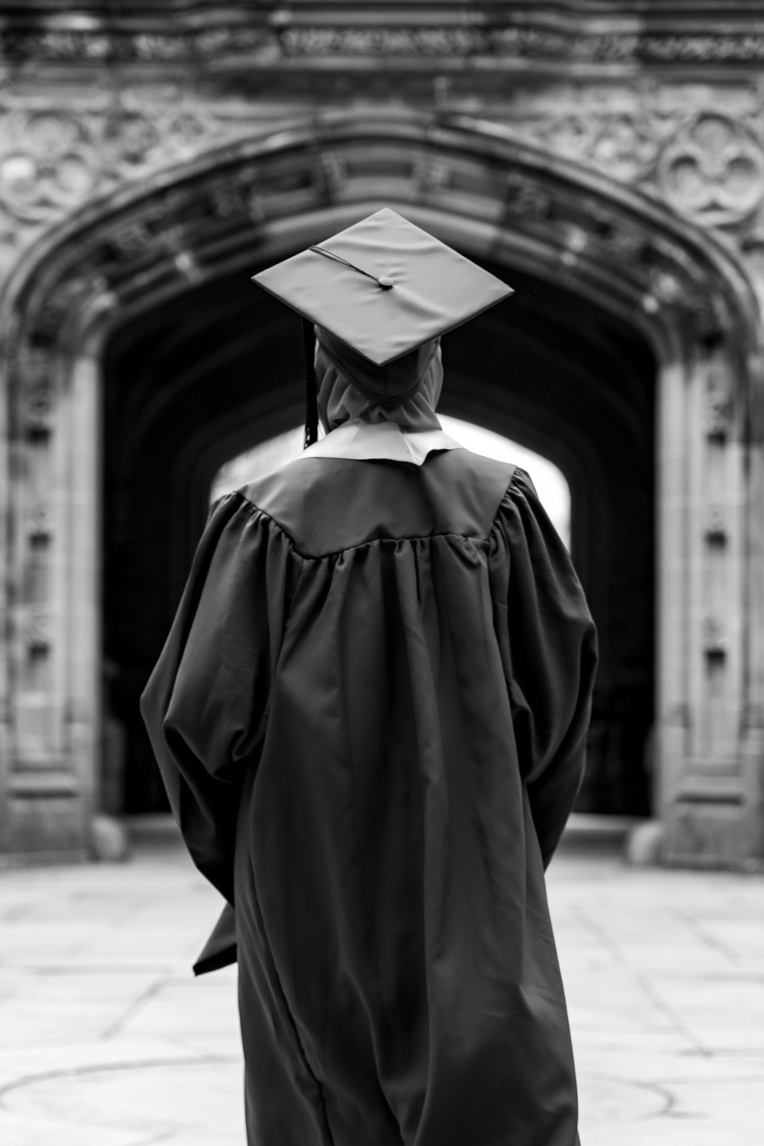 A graduate in cap and gown stands facing an ornate stone archway in black and white photography.