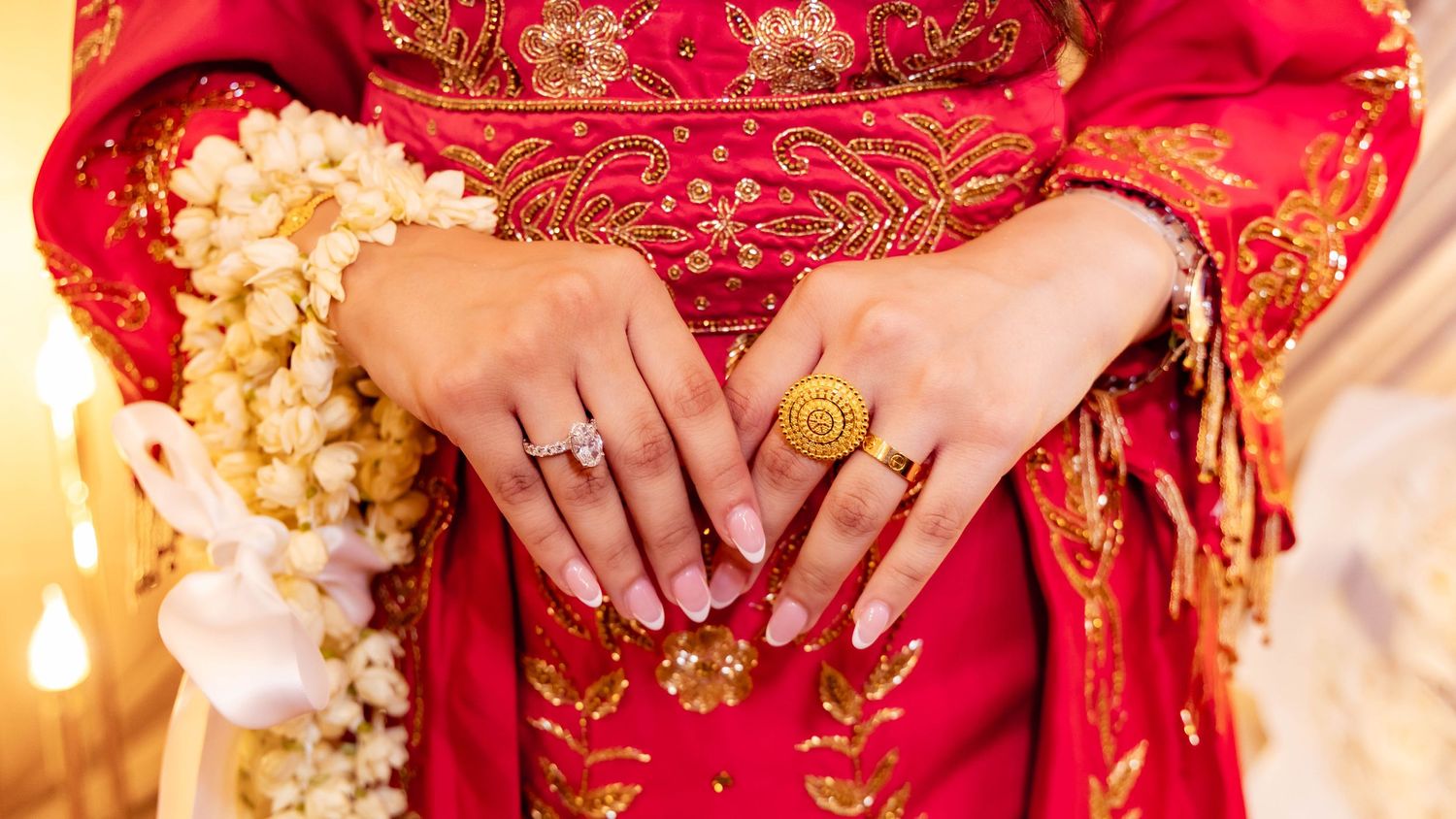 Close up of hands wearing traditional gold rings and jewelry against a red and gold embroidered qipao dress.
