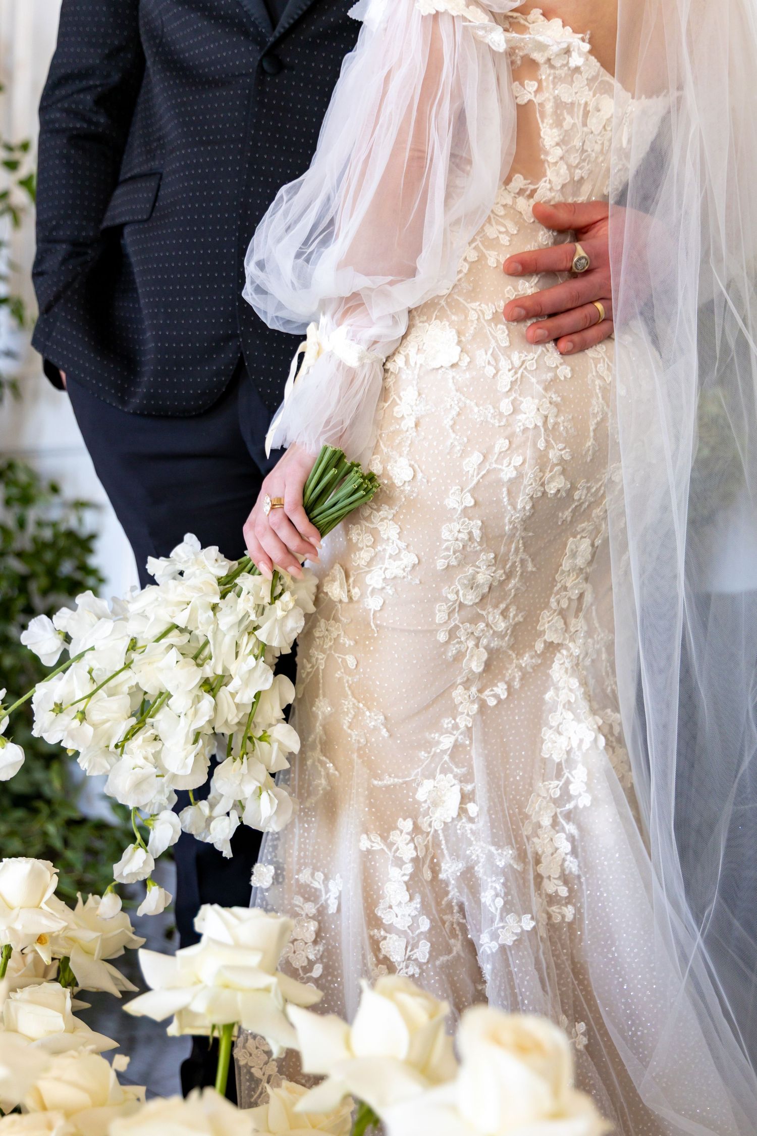 Close-up of bride in lace dress holding white bouquet next to groom in dark suit.