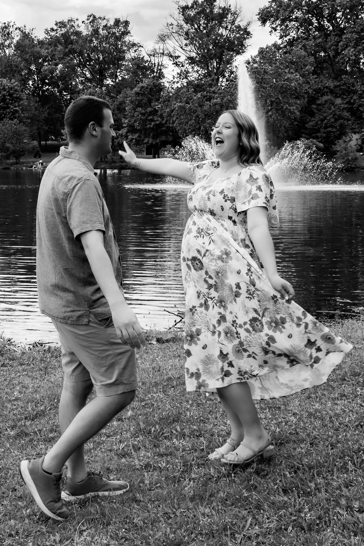 Couple playfully poses by a lake with fountain, woman wearing floral dress dancing in black and white photo.