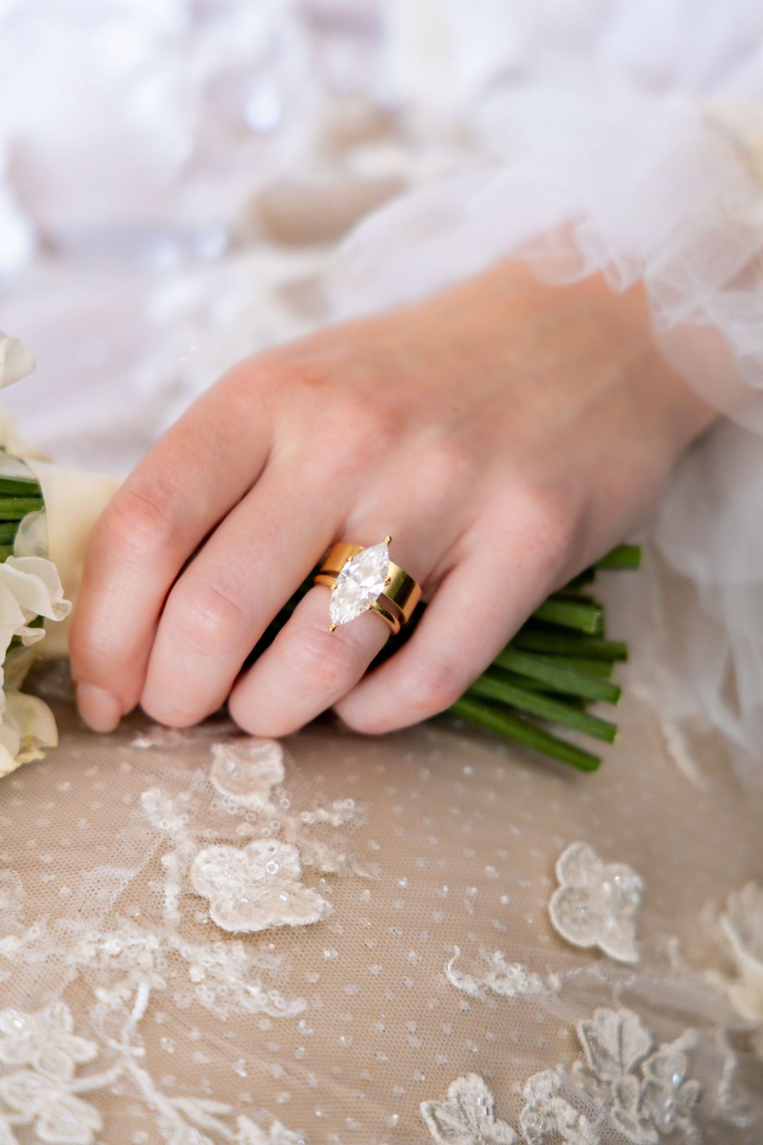 A hand wearing a gold engagement ring holds a white wedding bouquet against delicate lace fabric.