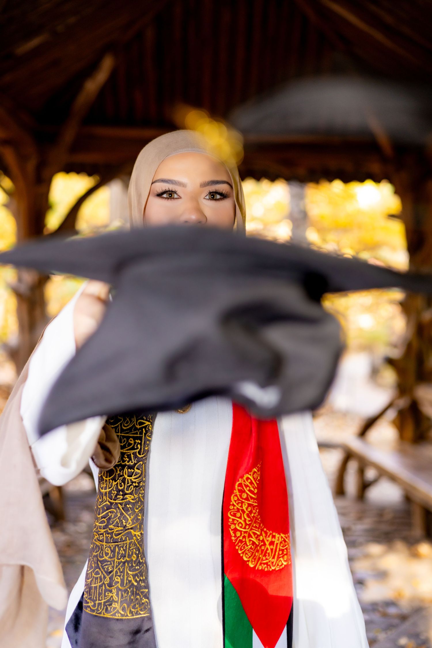 A graduate in traditional dress holding up a graduation cap against a warm glowing background. Graduation portraits.