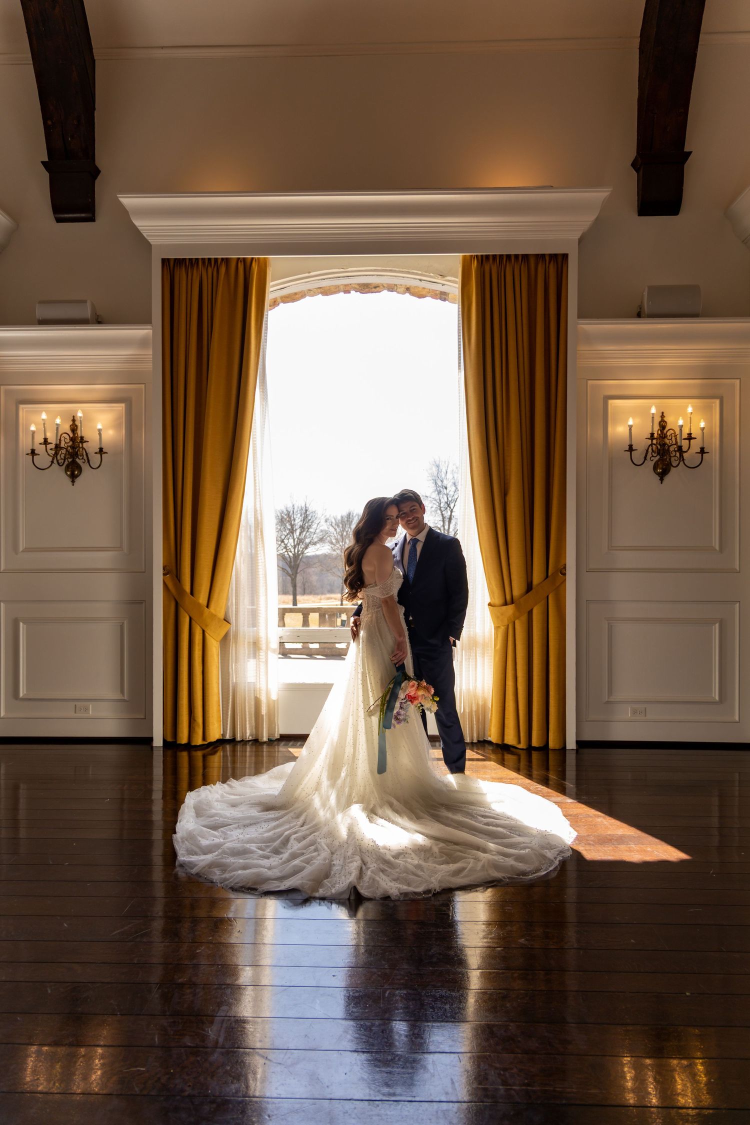 A wedding couple silhouetted against bright window light in an elegant ballroom with gold curtains and crystal sconces.