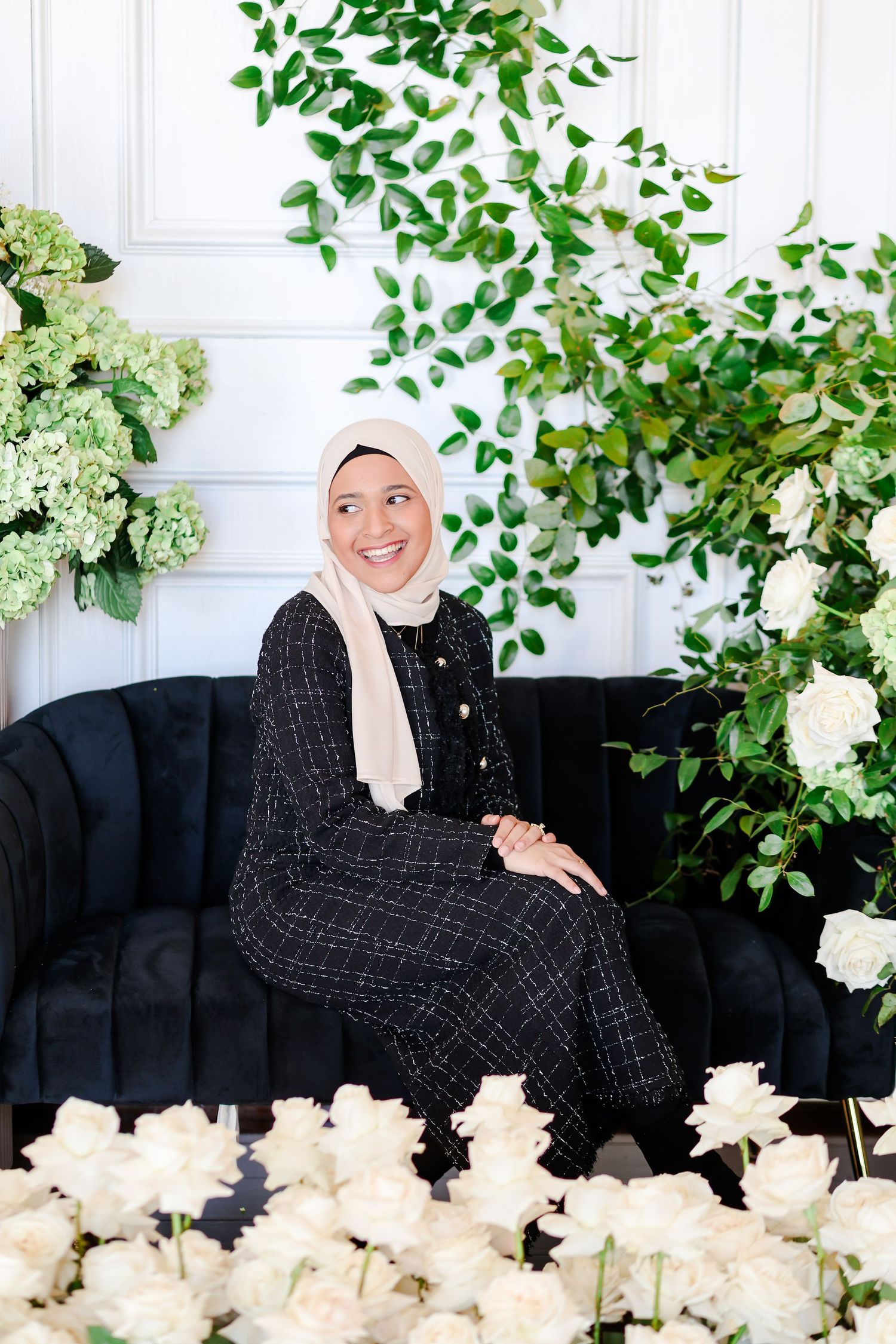Elegant floral photoshoot setting with a black velvet couch, greenery and white hydrangeas against a white paneled wall.