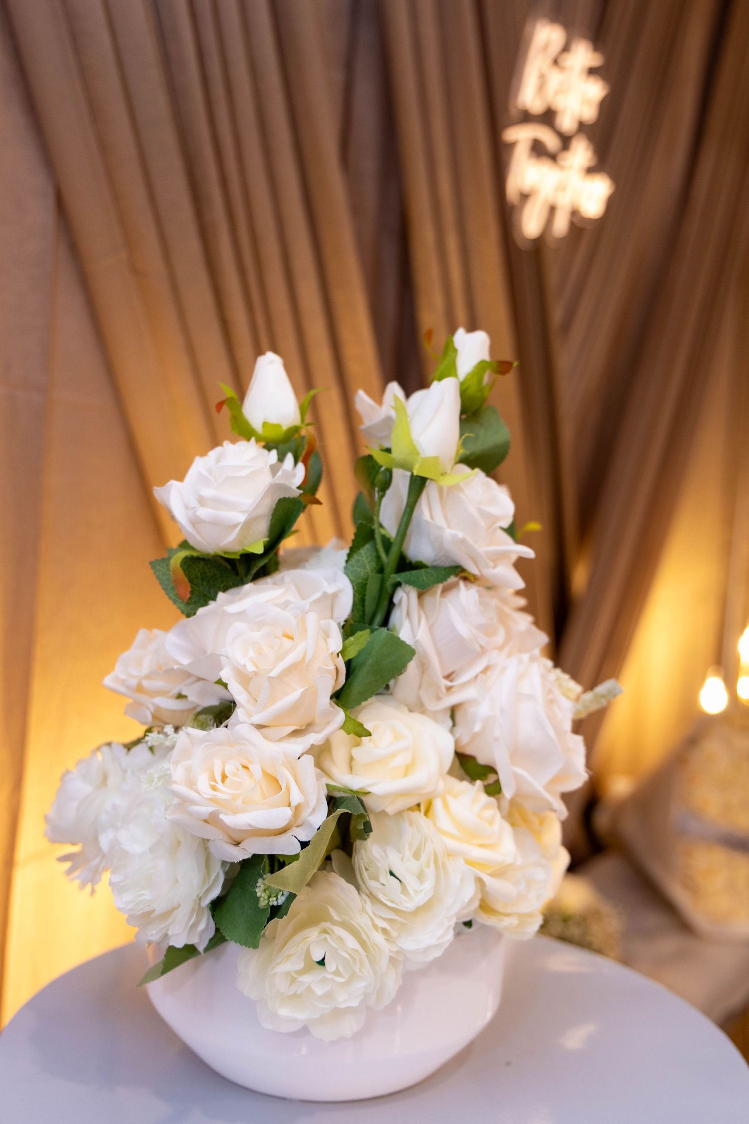 A white floral centerpiece arrangement of cream roses in a round vase against elegant draped fabric with warm lighting.