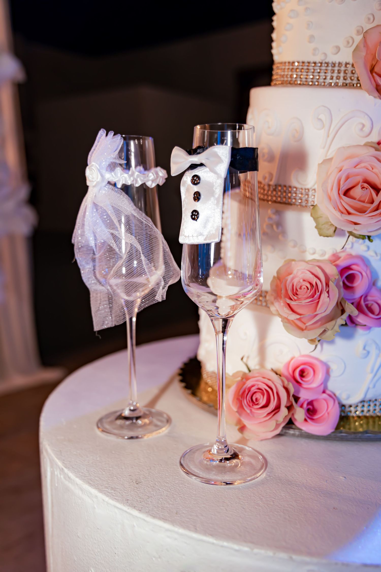 Bride and groom champagne glasses decorated with tulle veil and tuxedo design next to wedding cake.