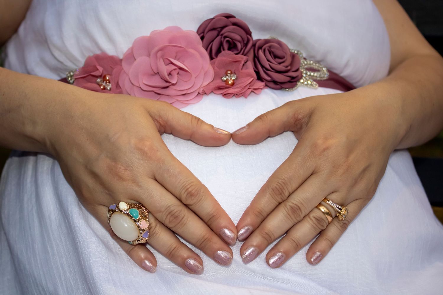 Hands forming a heart shape over a white dress with pink roses and decorative rings.