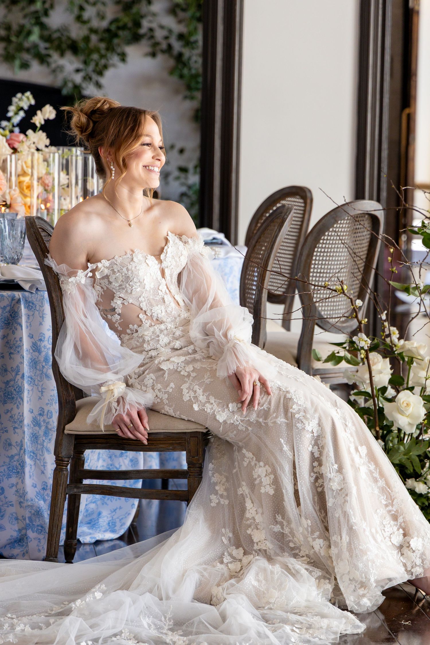 Bride in off-shoulder lace gown sits gracefully on chair surrounded by floral arrangements.